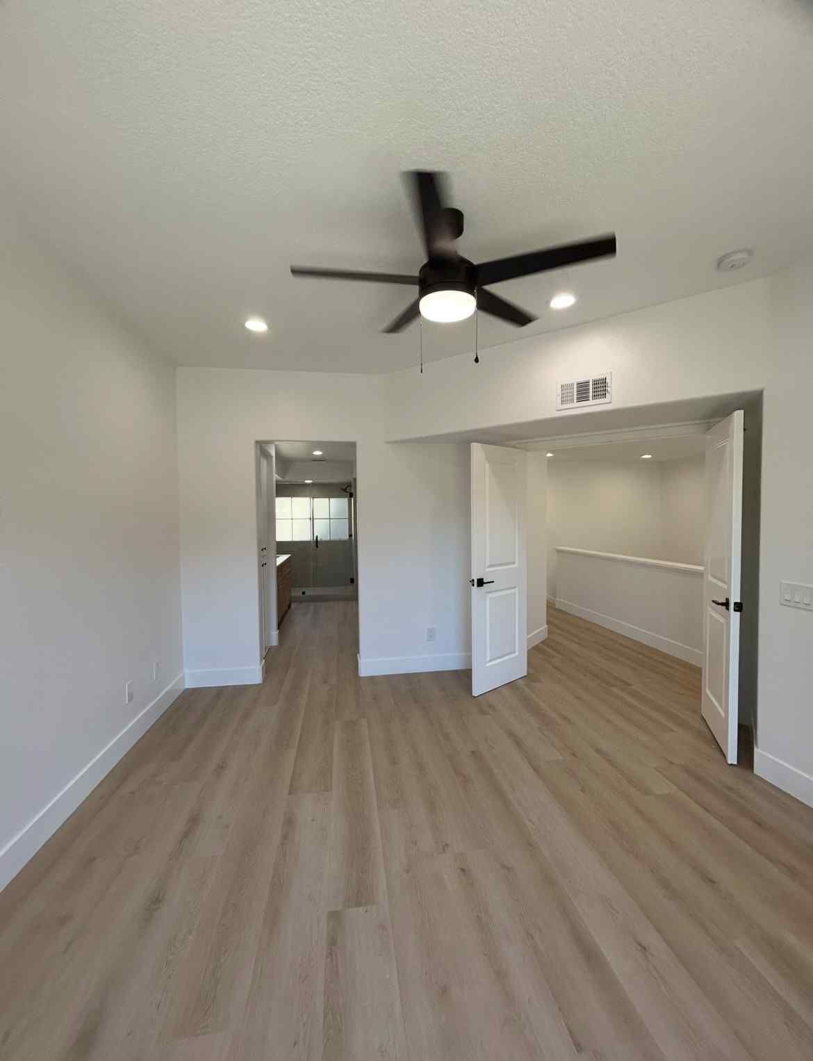 Empty modern room with light wood floors, white walls, ceiling fan, and open doorway to a hallway
