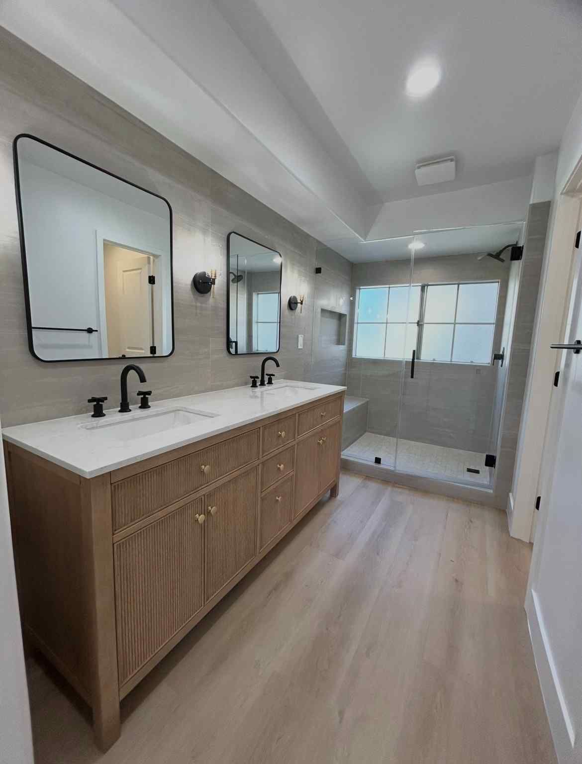 Modern bathroom with long wood vanity, two mirrors, black faucets, and a walk-in shower with frosted glass.