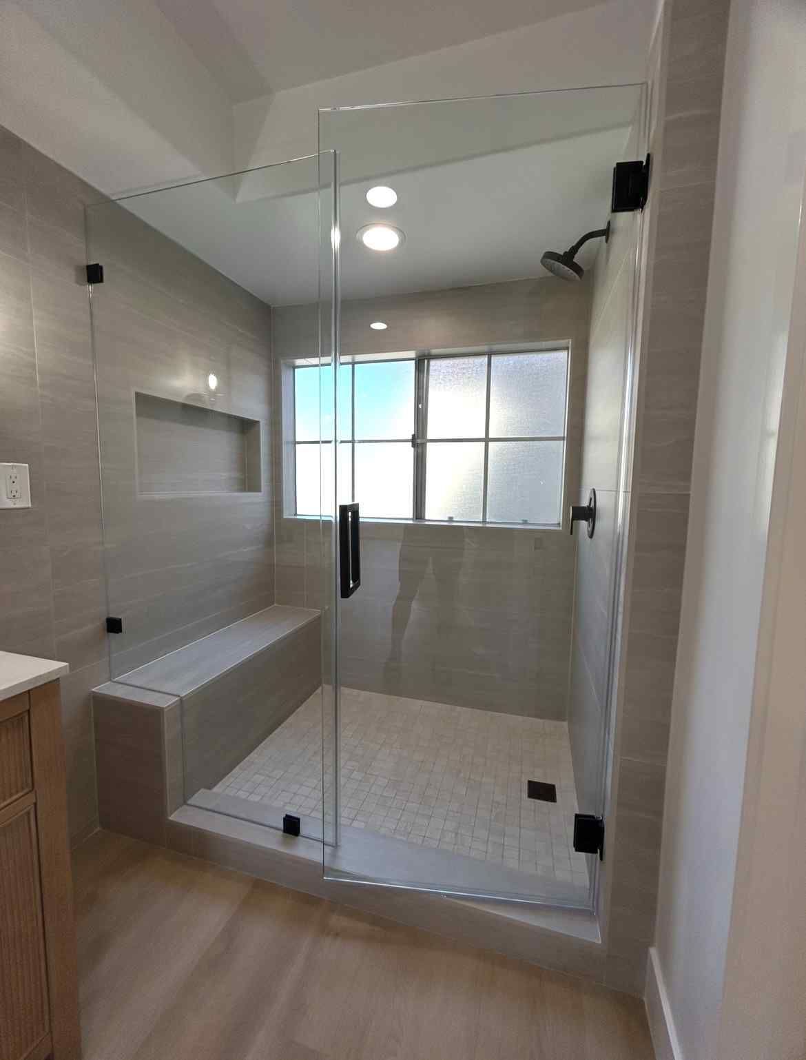 Modern bathroom with glass shower enclosure, gray tile, bench, and frosted window