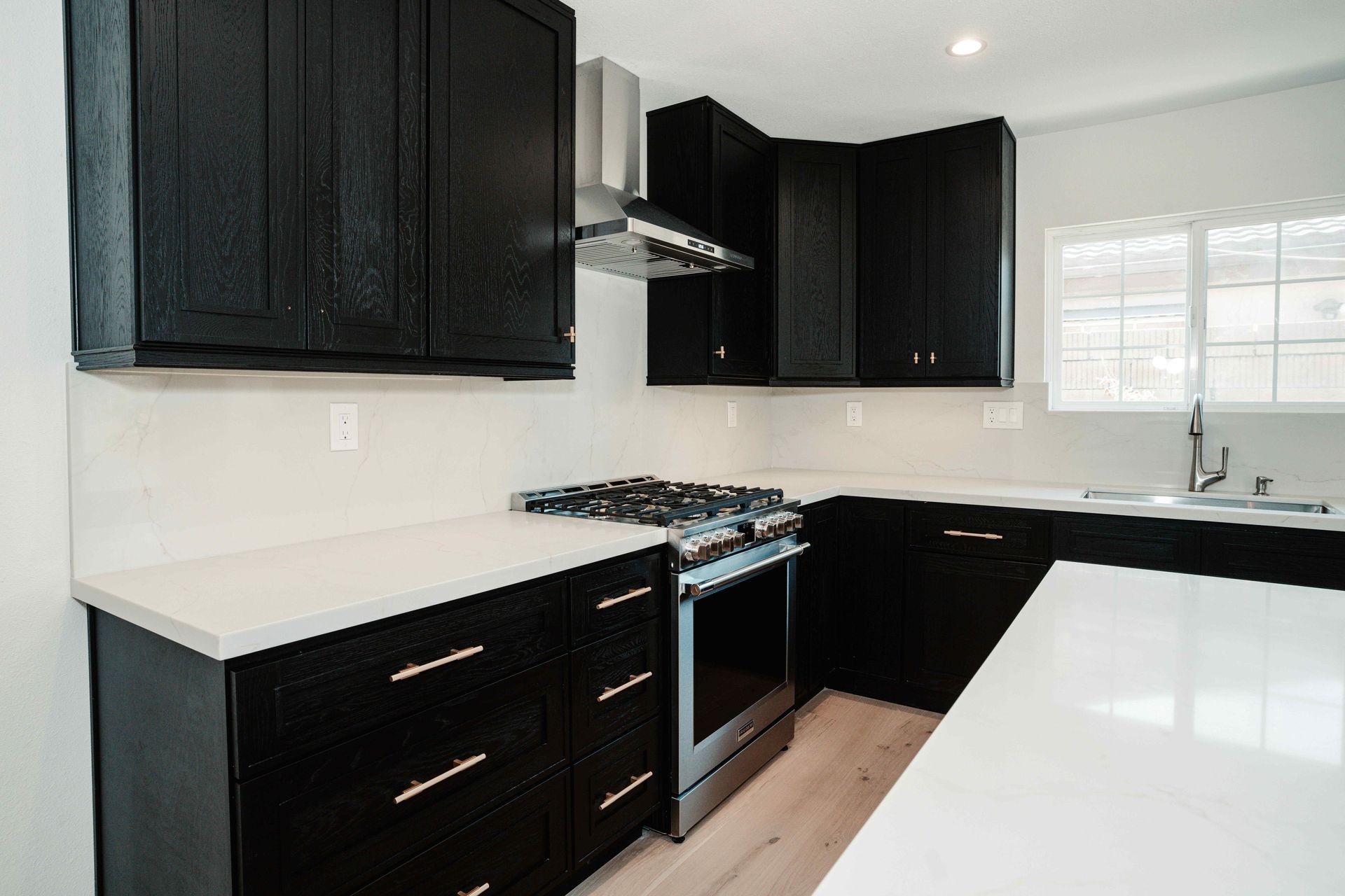 Modern kitchen with black cabinets, white countertops, gas stove, and window over the sink.