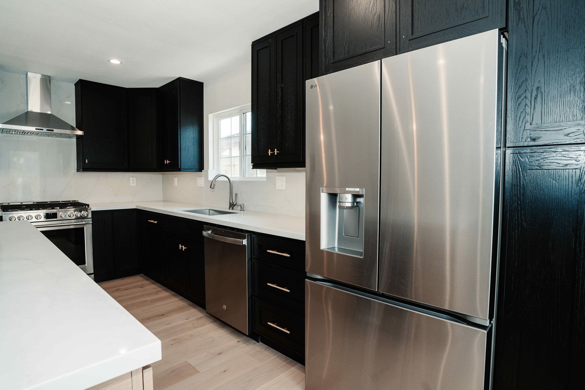 Modern kitchen with black cabinets, stainless steel fridge, white countertops, and light wood flooring