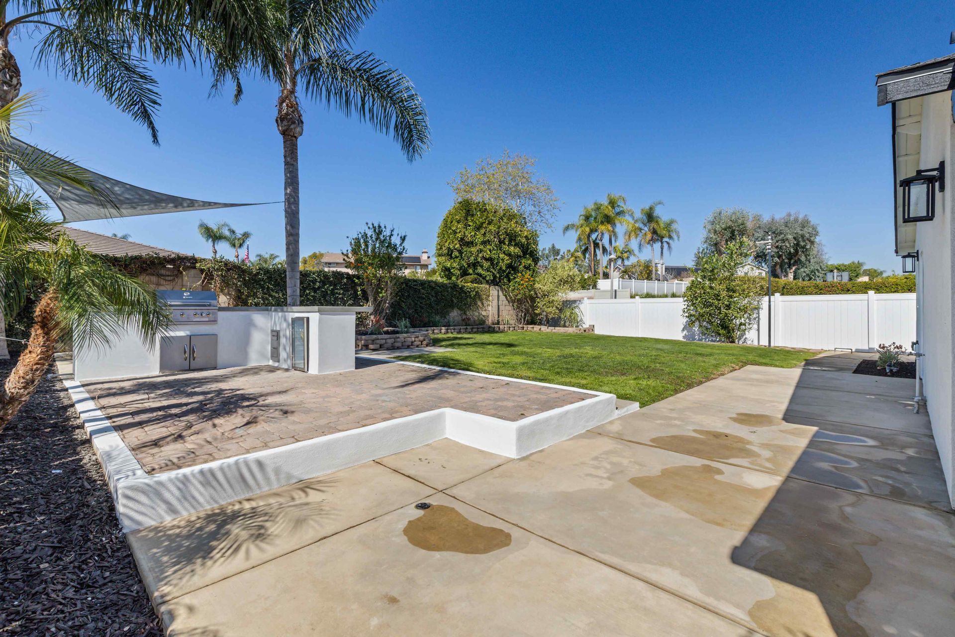 Backyard with concrete patio, built-in grill, grassy area, white fence, and palm trees under a blue sky.
