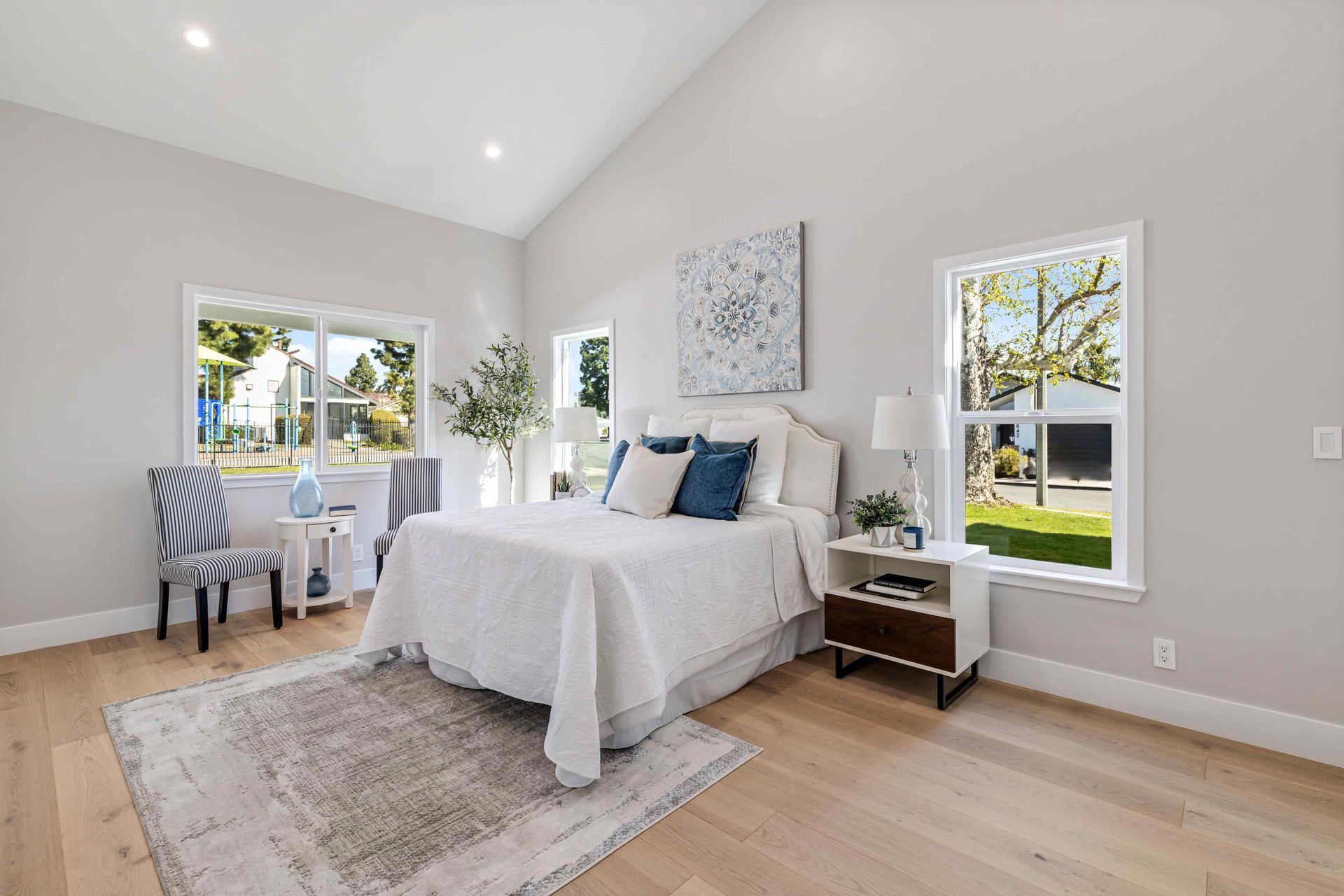Bright bedroom with vaulted ceiling, white bed, rug, desk, and two windows with a tree view.