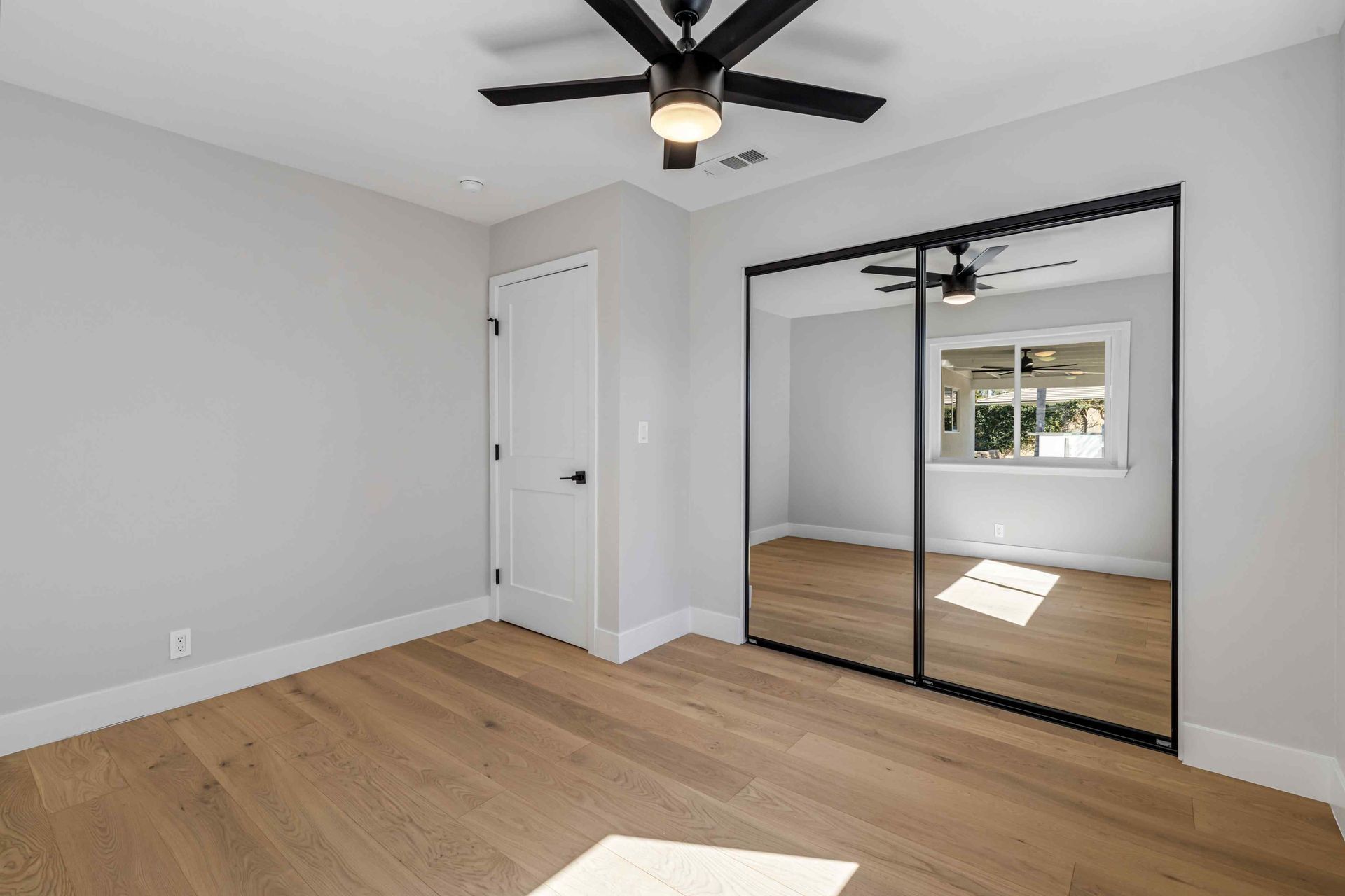 Empty bedroom with light wood floors, gray walls, mirrored closet doors, and a ceiling fan.