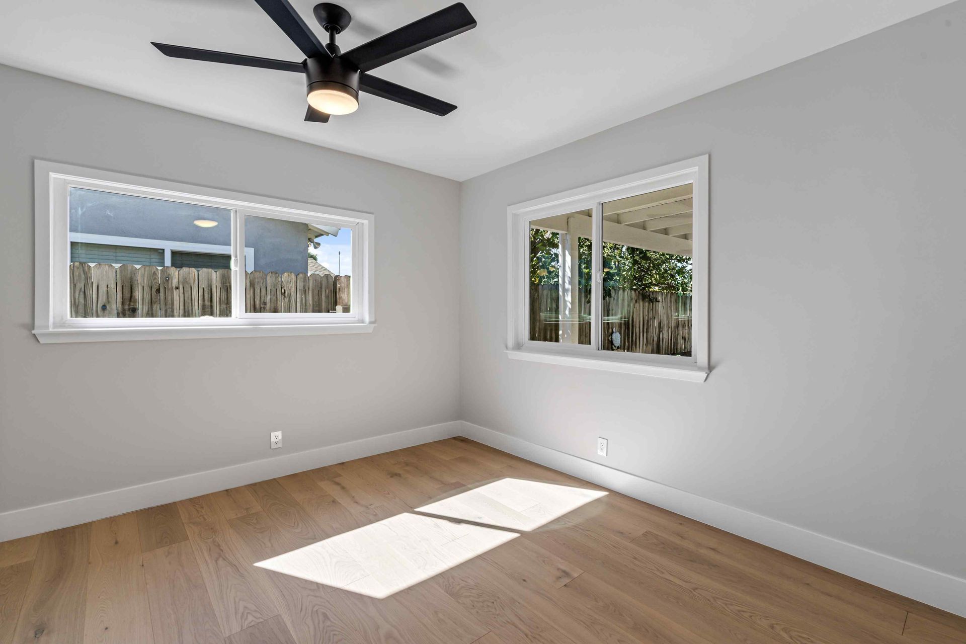Empty bedroom with light wood floors, gray walls, and two windows. A black ceiling fan hangs overhead.