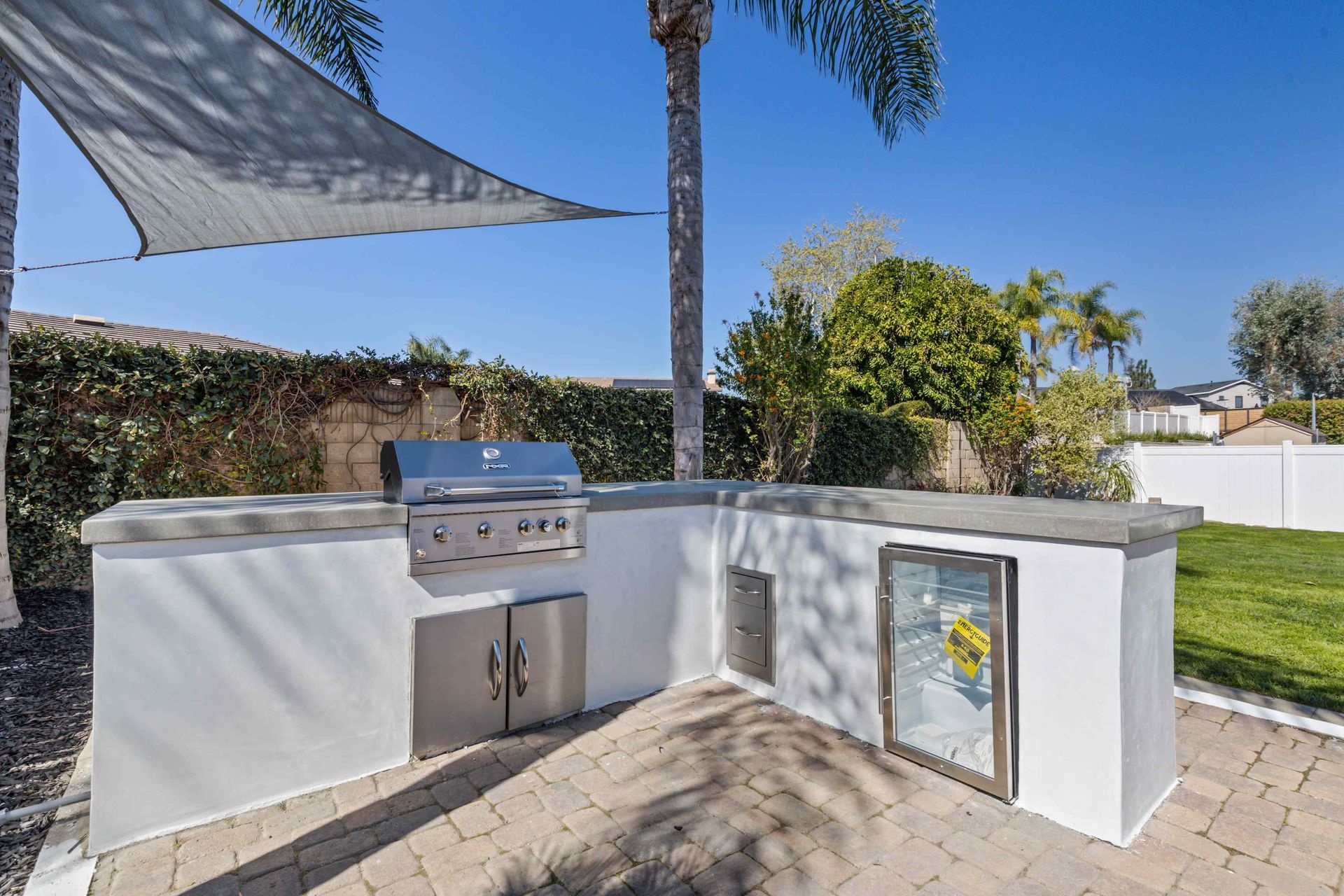 Outdoor kitchen with grill, refrigerator, and countertop, on a brick patio, under a sun shade.