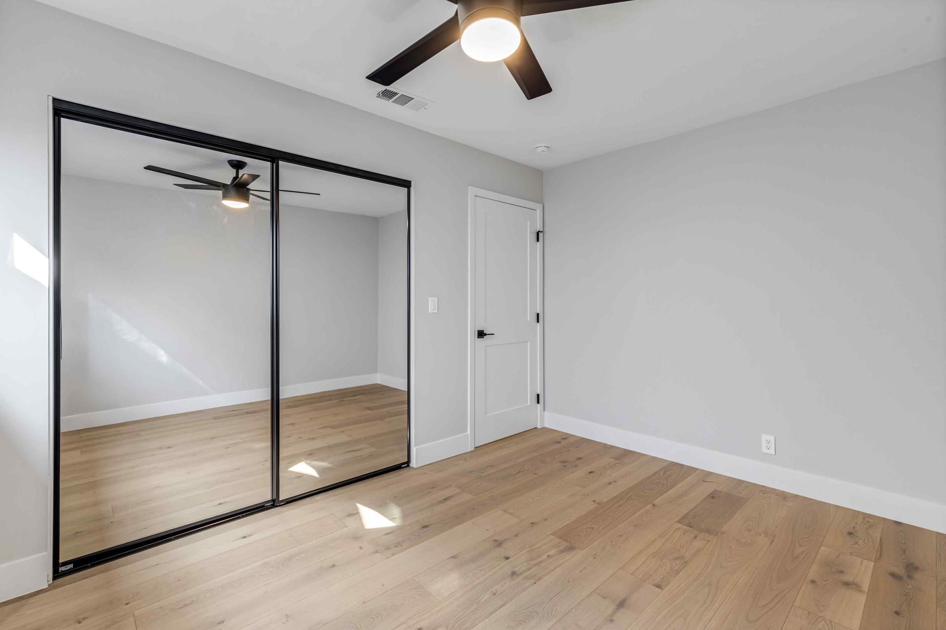 Empty bedroom with hardwood floors, large mirrored closet doors, white door, and black ceiling fan.