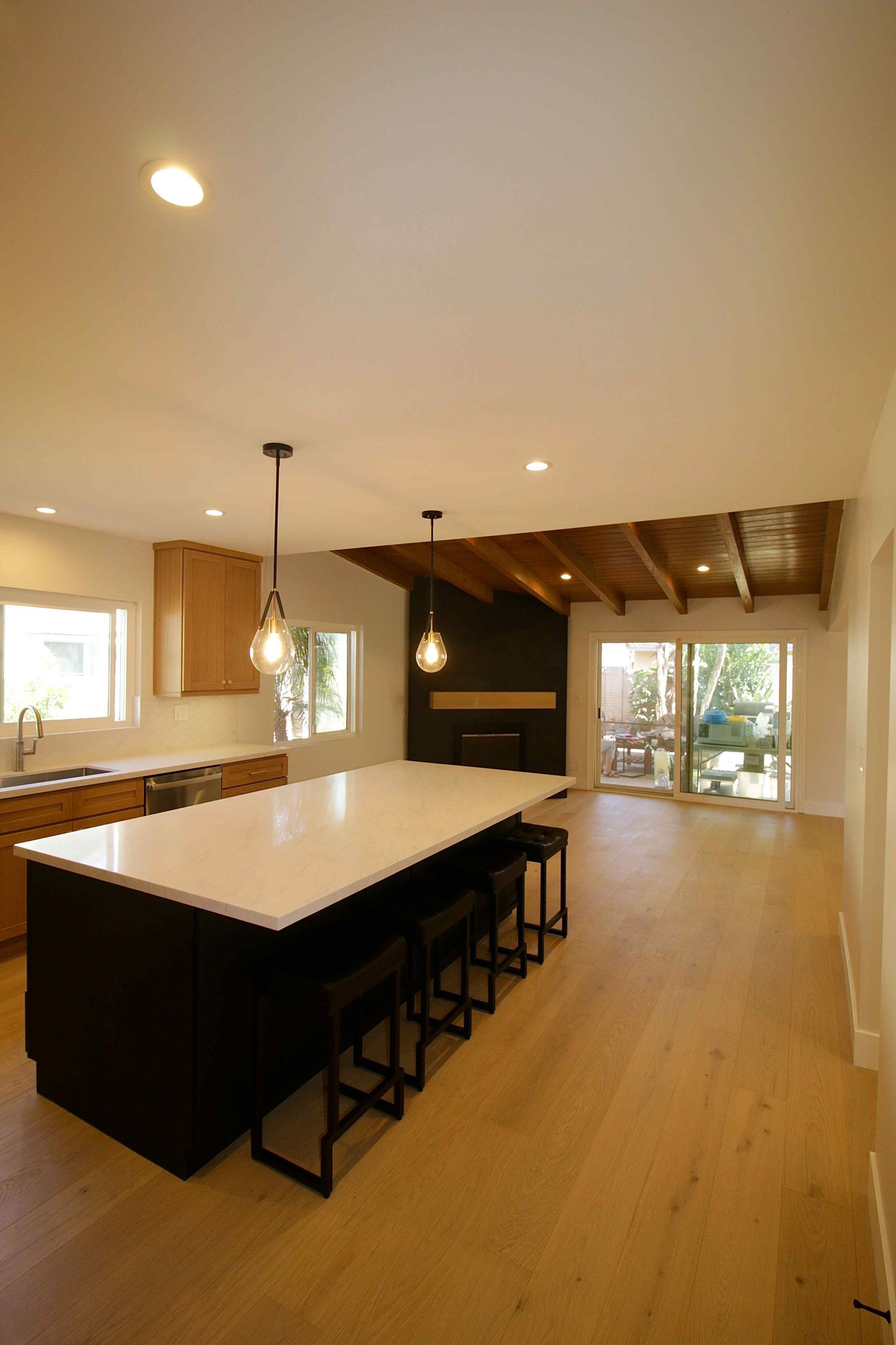 Modern kitchen with large island, wood floors, pendant lights, and open living area with fireplace and windows