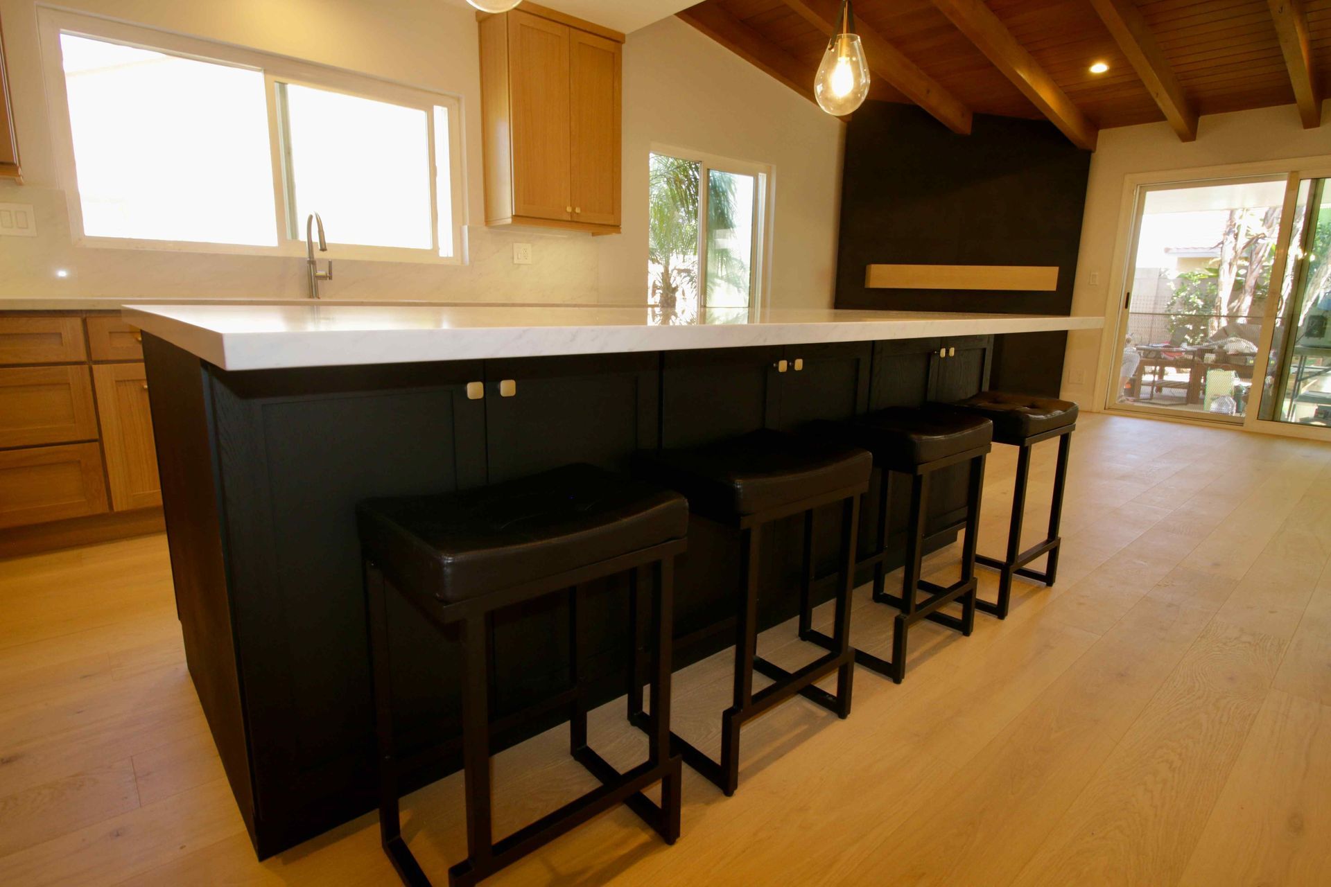 Modern kitchen with dark island and four stools, wood floors, large windows, and pendant lights
