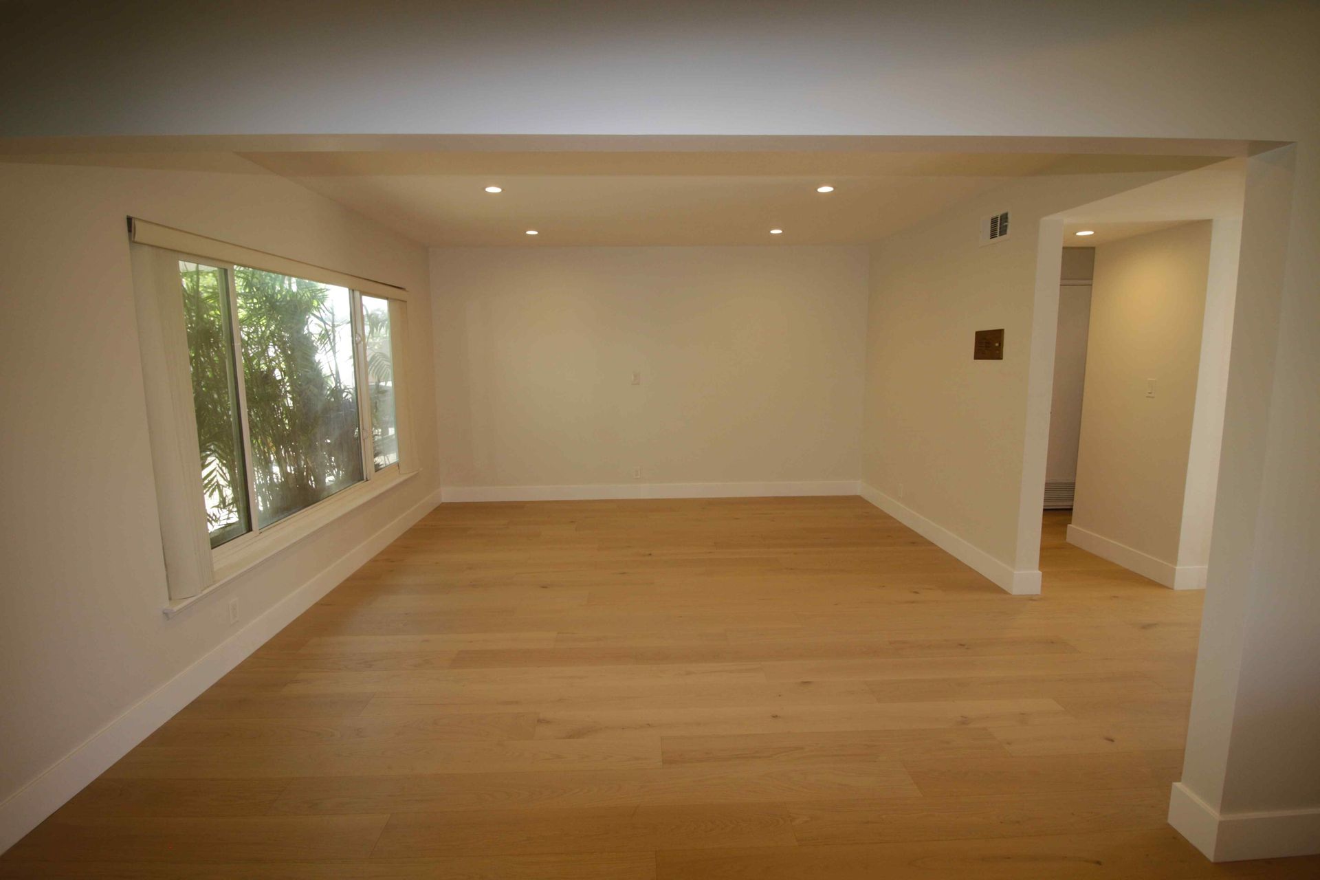 Empty modern living room with hardwood floors, white walls, and large windows on the left.