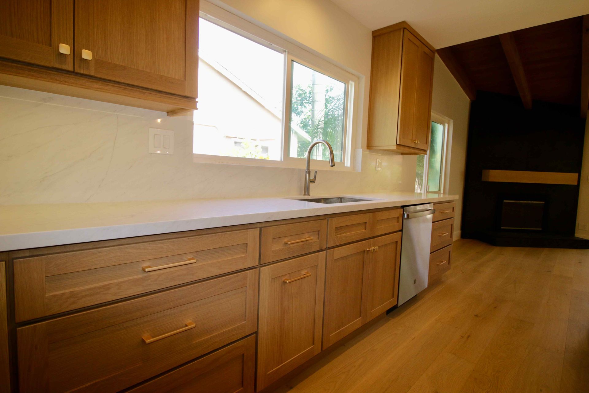 Bright wood kitchen with white countertops, sink under a window, and dark vaulted dining area beyond