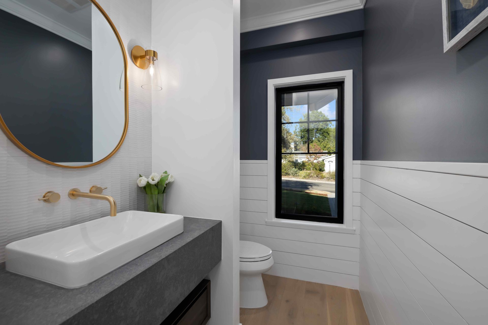 Powder room with white and navy walls, gold fixtures, rectangular sink, oval mirror, and small window.
