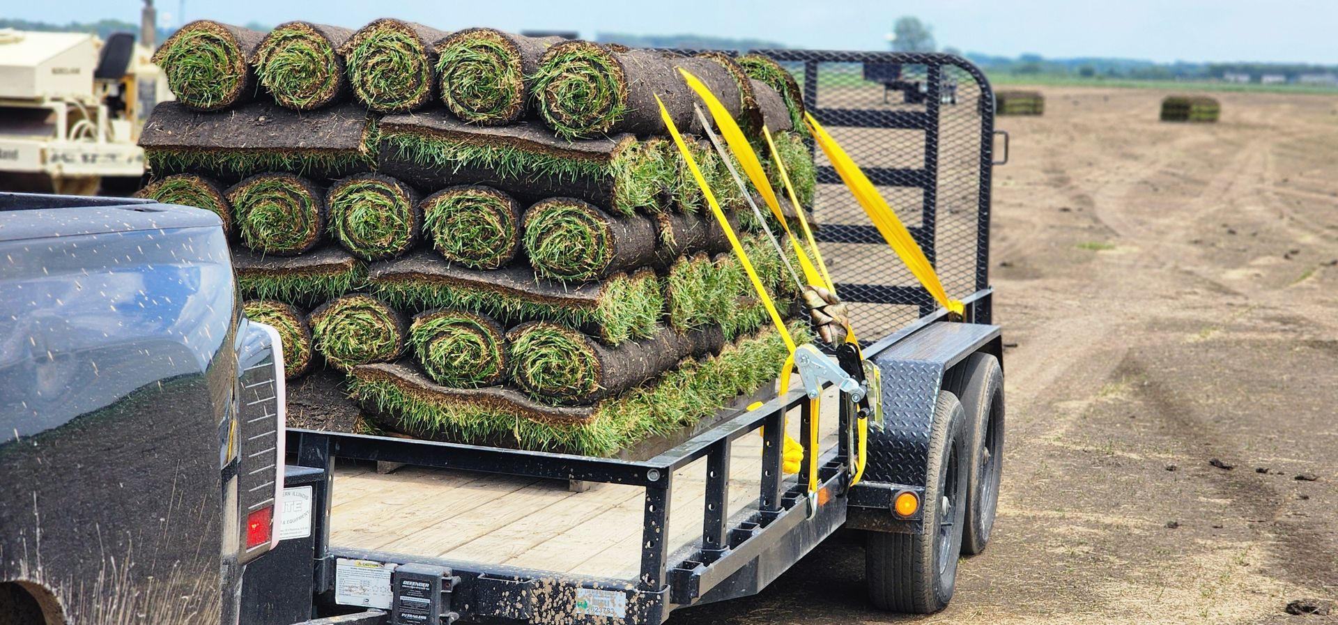 A trailer loaded with rolled sod being hauled down a dirt road. Bright yellow straps secure the load.