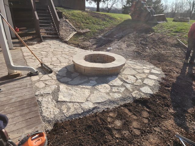 Stone fire pit on a stone patio, dirt ground, steps and grassy hill in the background.