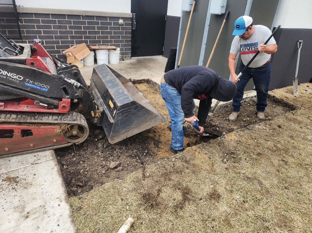 Two people digging near a small excavator. One uses a shovel, the other bends over, working in an excavated area.