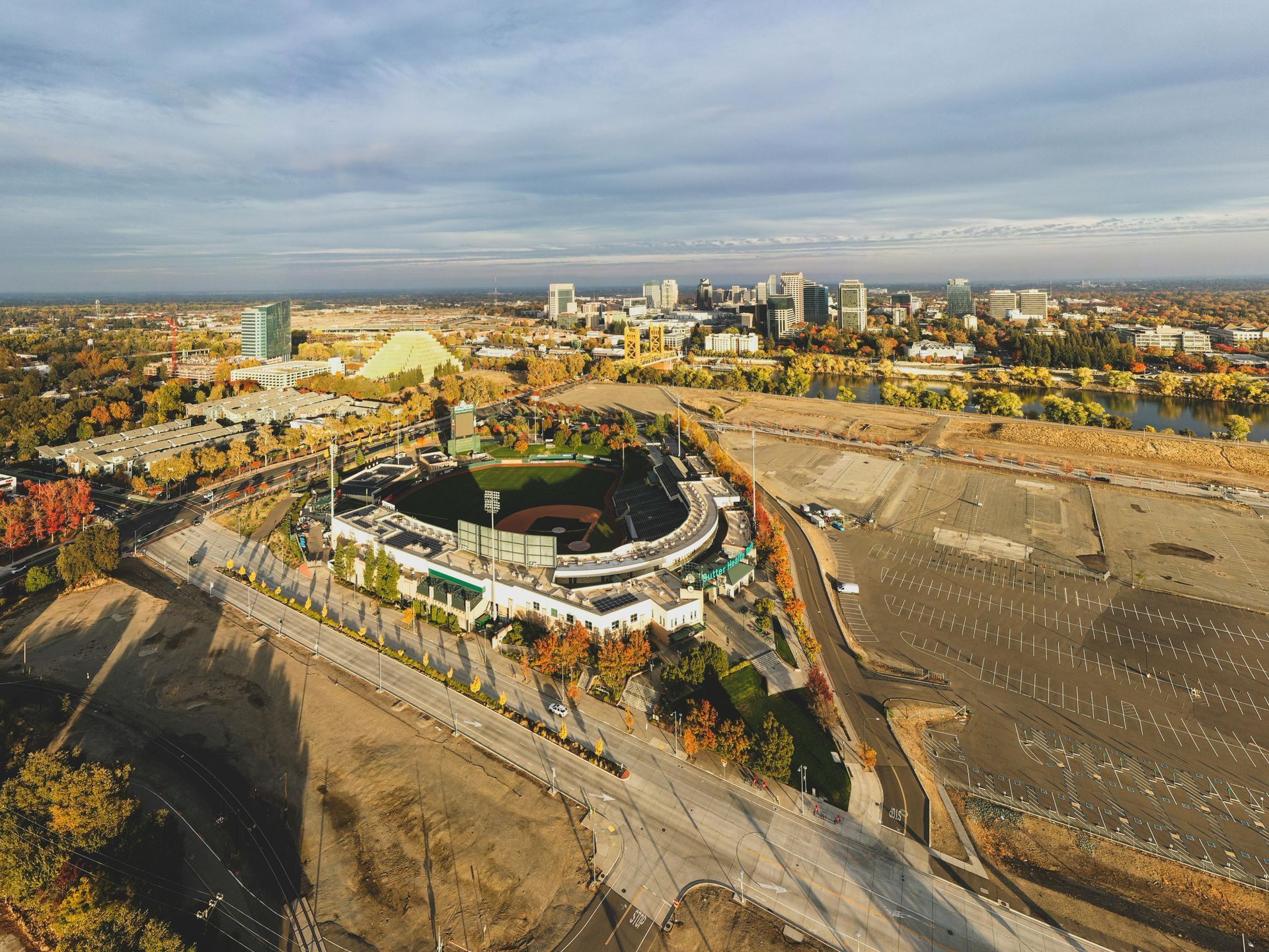 Aerial view of baseball stadium in city. Open parking lot, trees, and buildings visible under a cloudy sky.