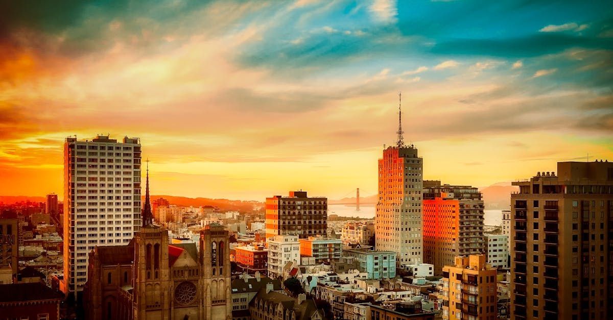 City skyline at sunset with colorful sky. Buildings silhouetted against vibrant orange and gold hues.