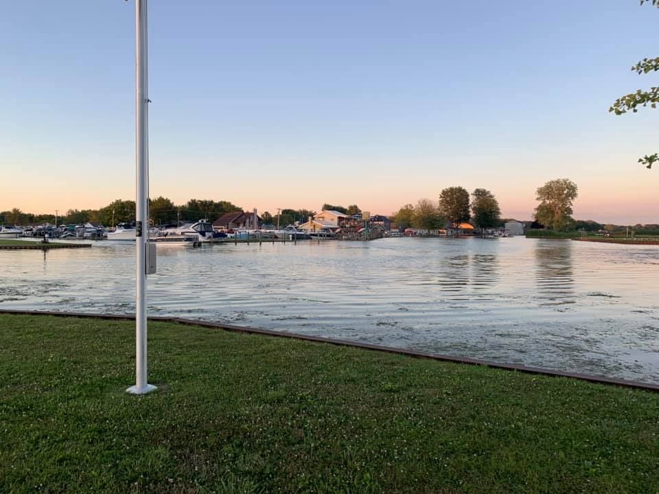 Flooded waterfront at sunset with calm water, grassy foreground, and a flagpole by the shore