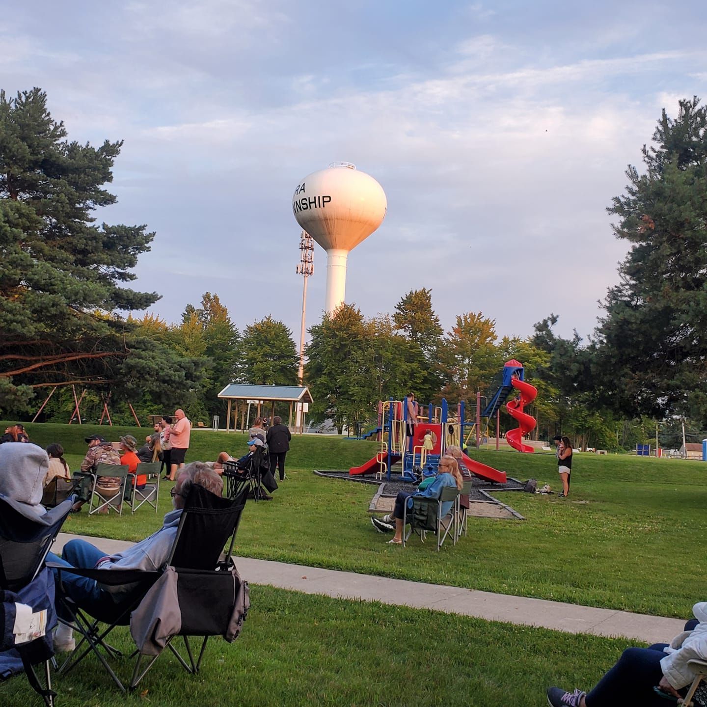 People at a park playground with a tall water tower and trees at sunset