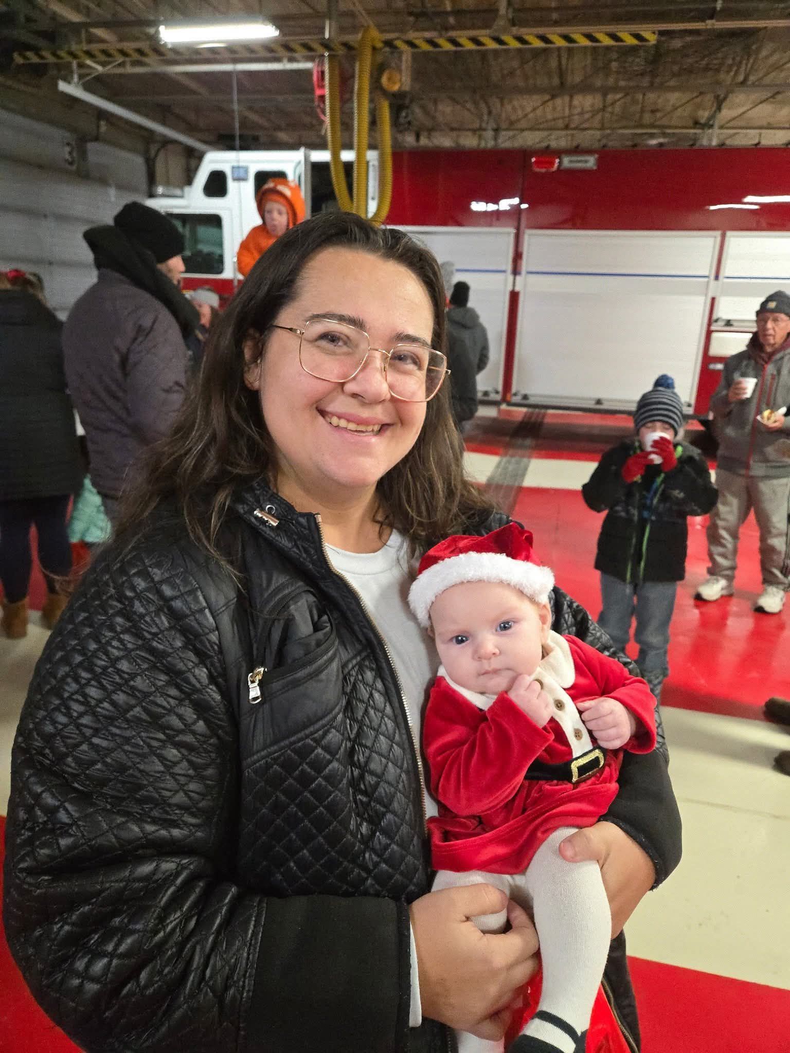 Smiling woman holding a baby in red holiday outfit inside a fire station with a fire truck behind them