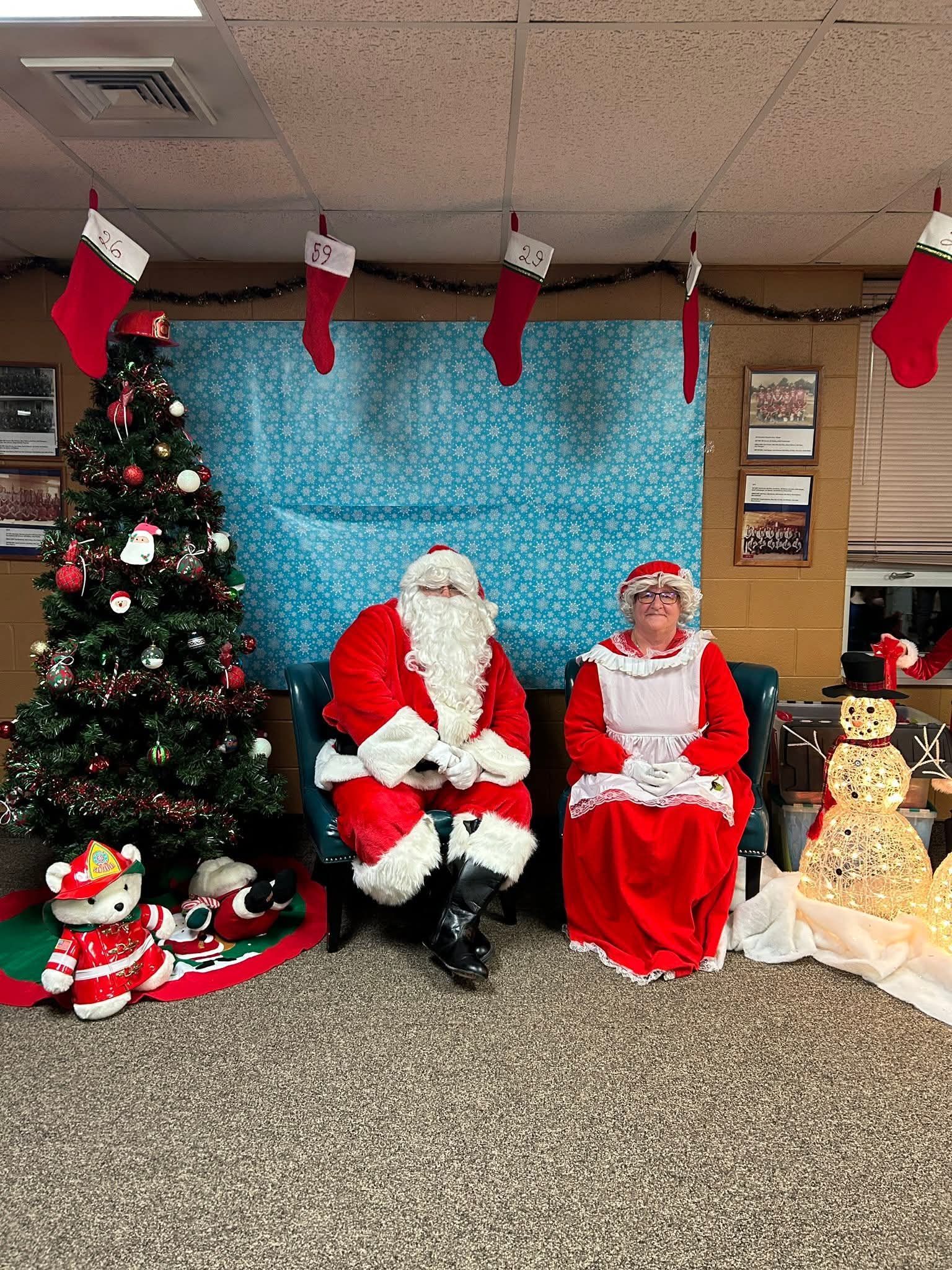 Santa and Mrs. Claus seated beside a decorated Christmas tree and holiday display at an indoor event