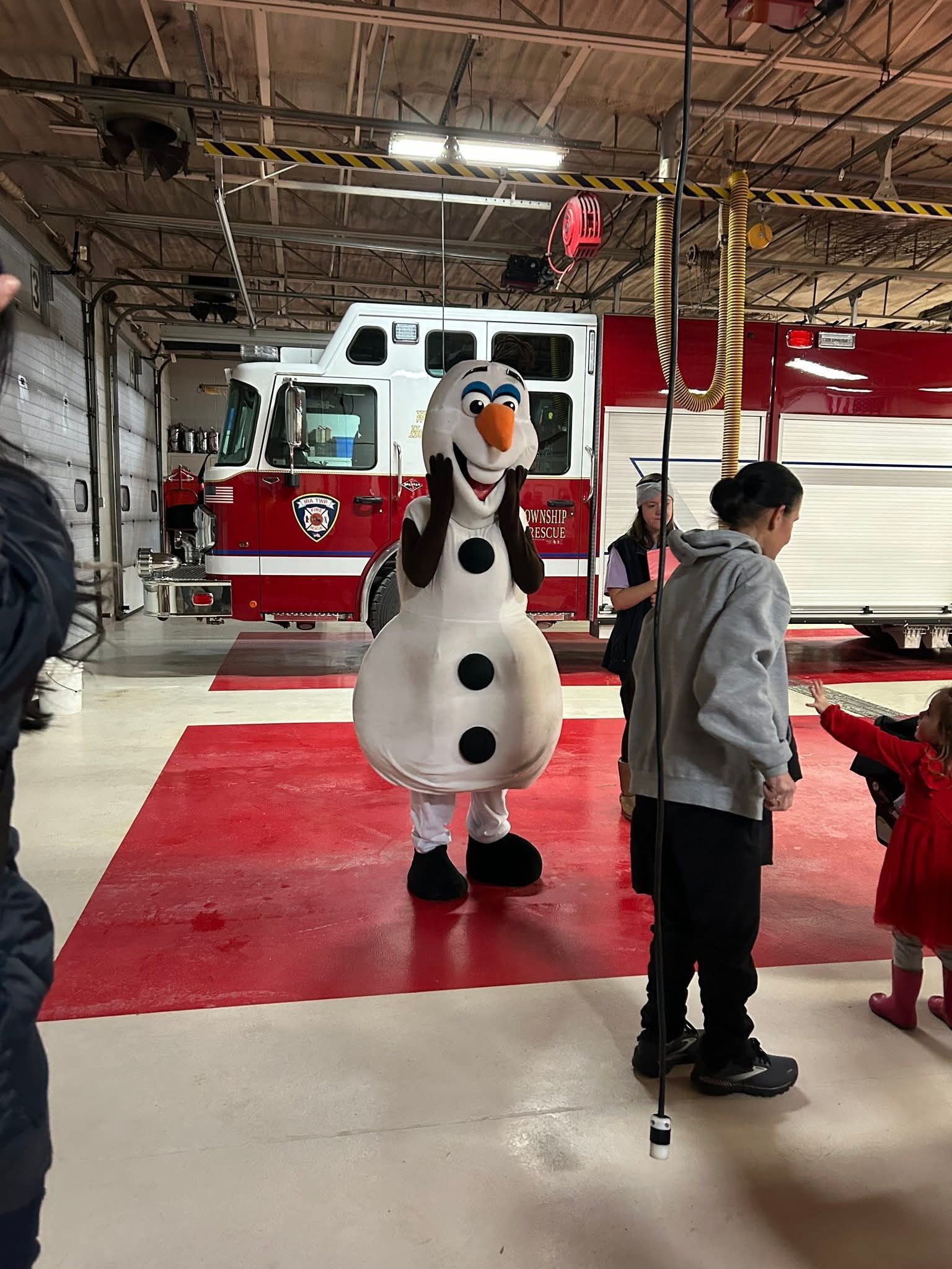 Olaf mascot in a fire station bay beside a red-and-white fire truck, with people nearby