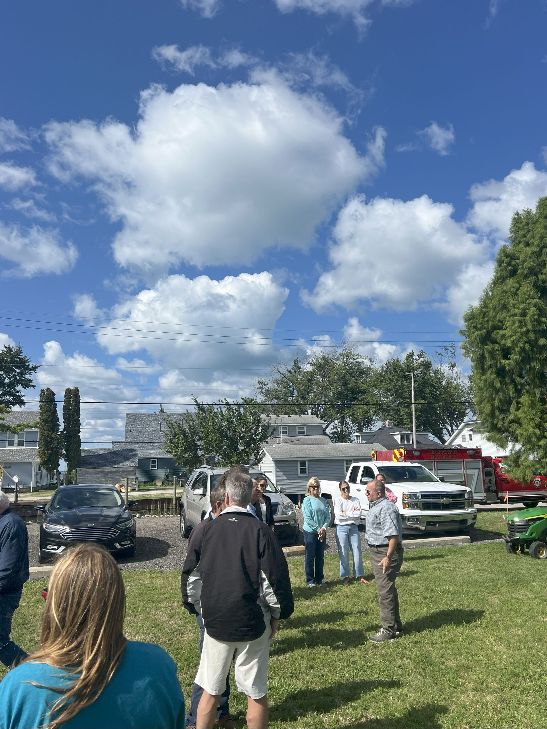 People standing on a grassy field under a blue sky with clouds, near parked cars and trees.