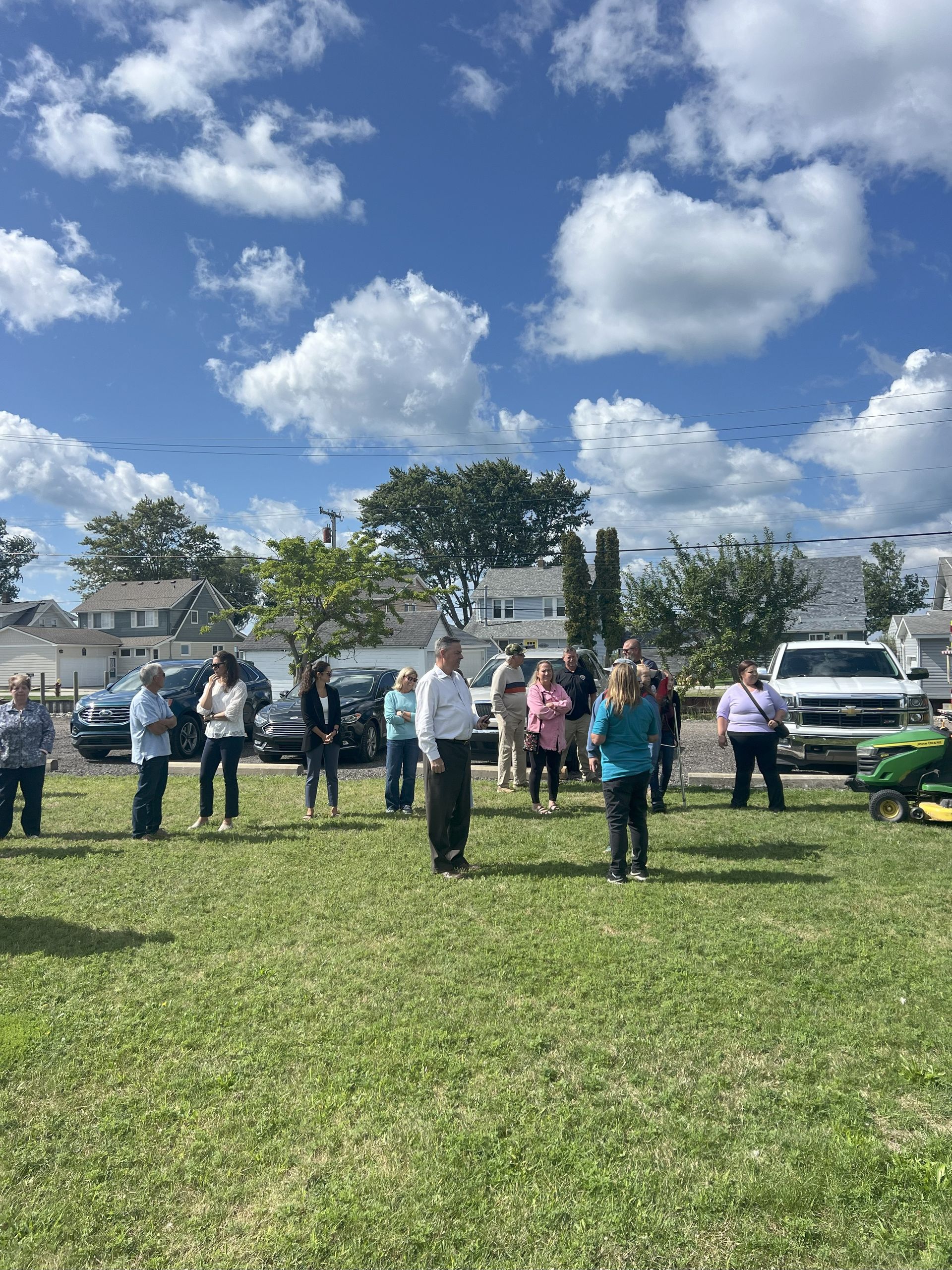 People standing on a grassy field under a bright blue sky with clouds, near parked cars and trees.