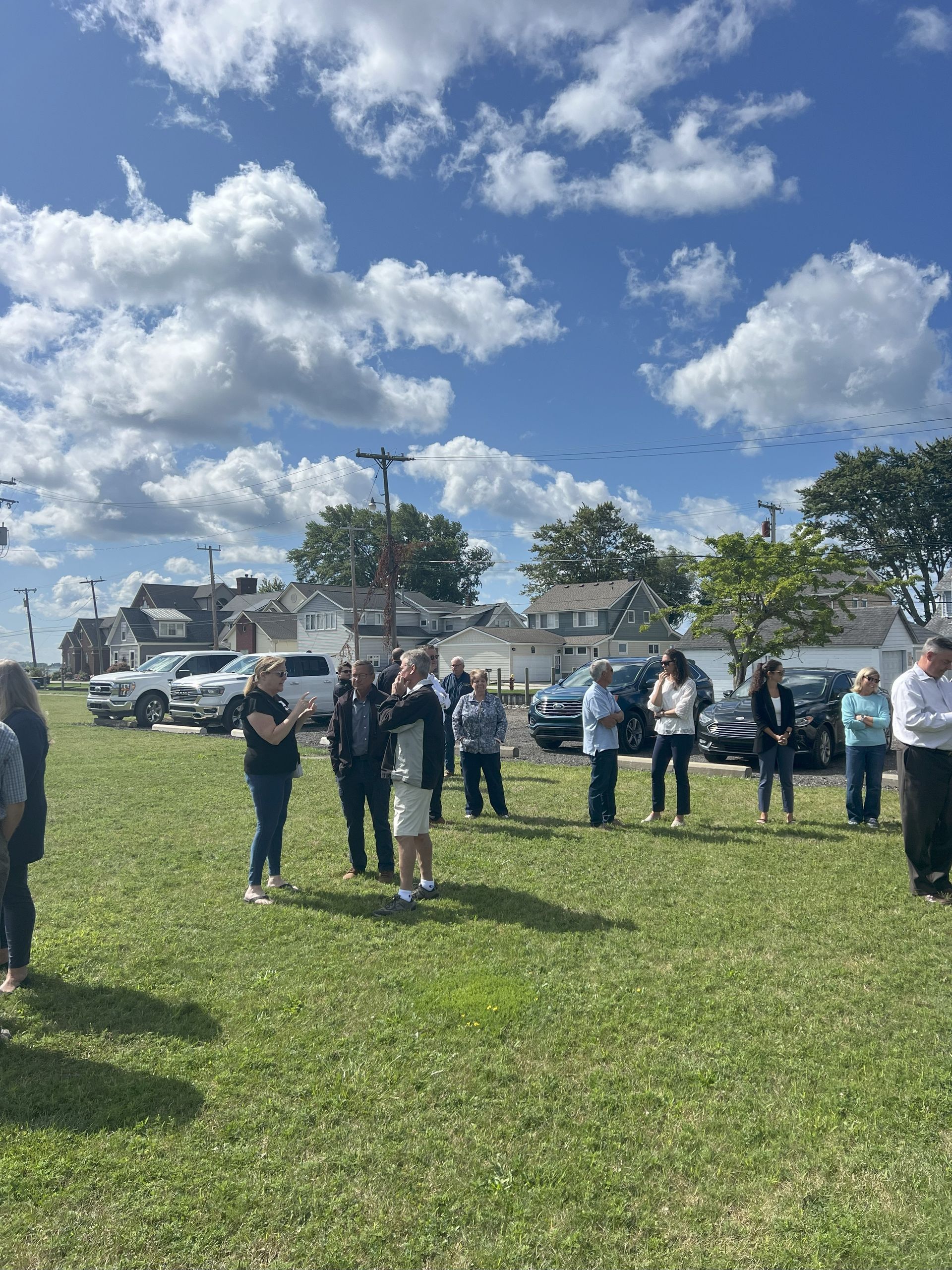 People standing on a grassy field under a blue sky with clouds, near parked cars and houses.