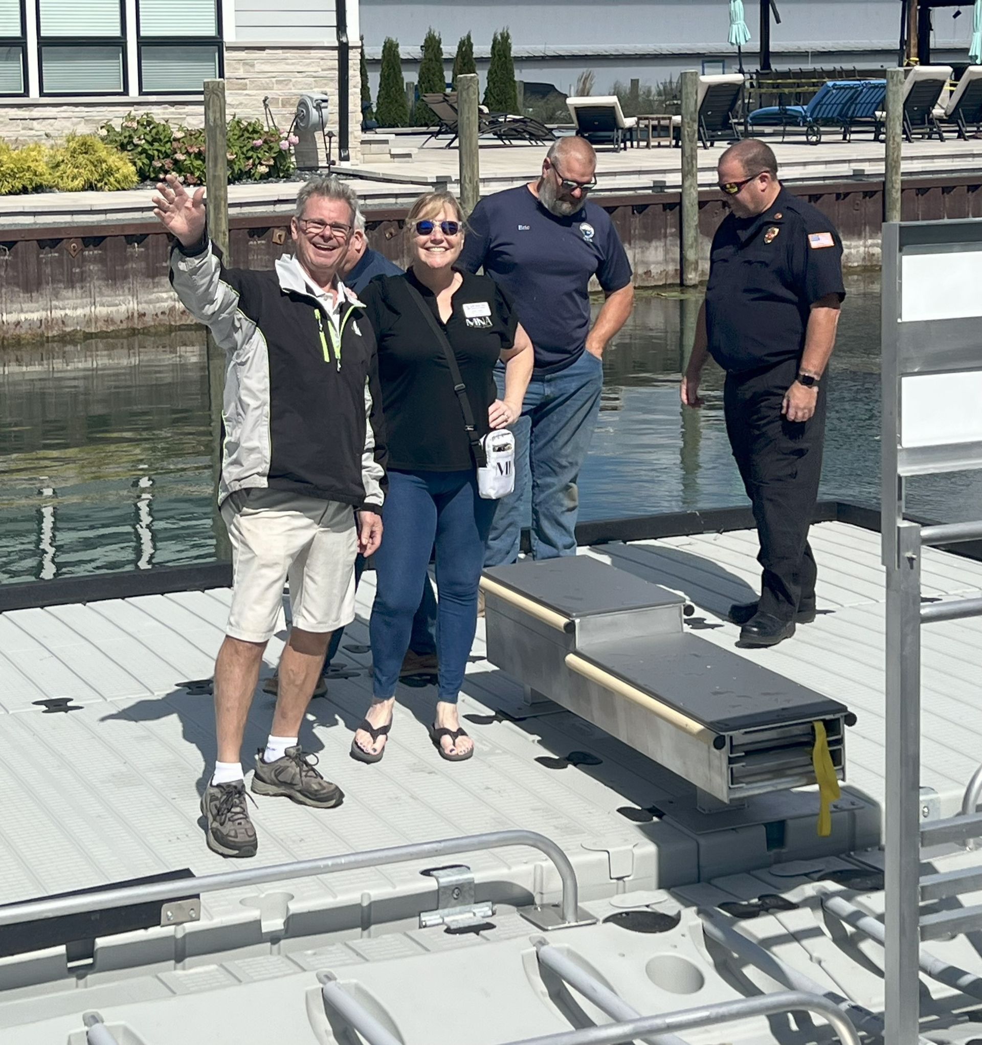 Four people standing on a dock by the water; two pose for a photo while one waves.