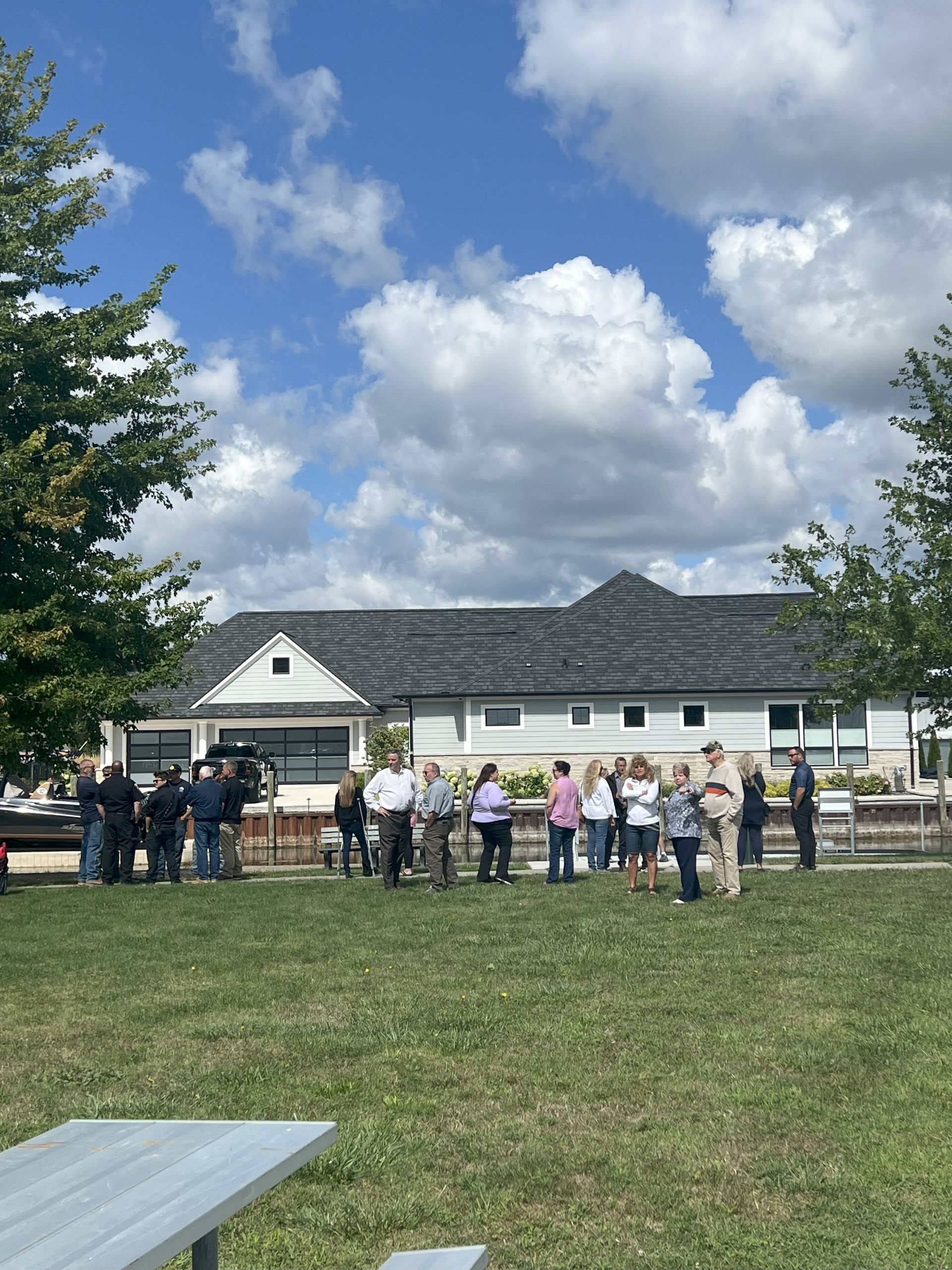 People gathered on a grassy field near a white building under a cloudy blue sky.