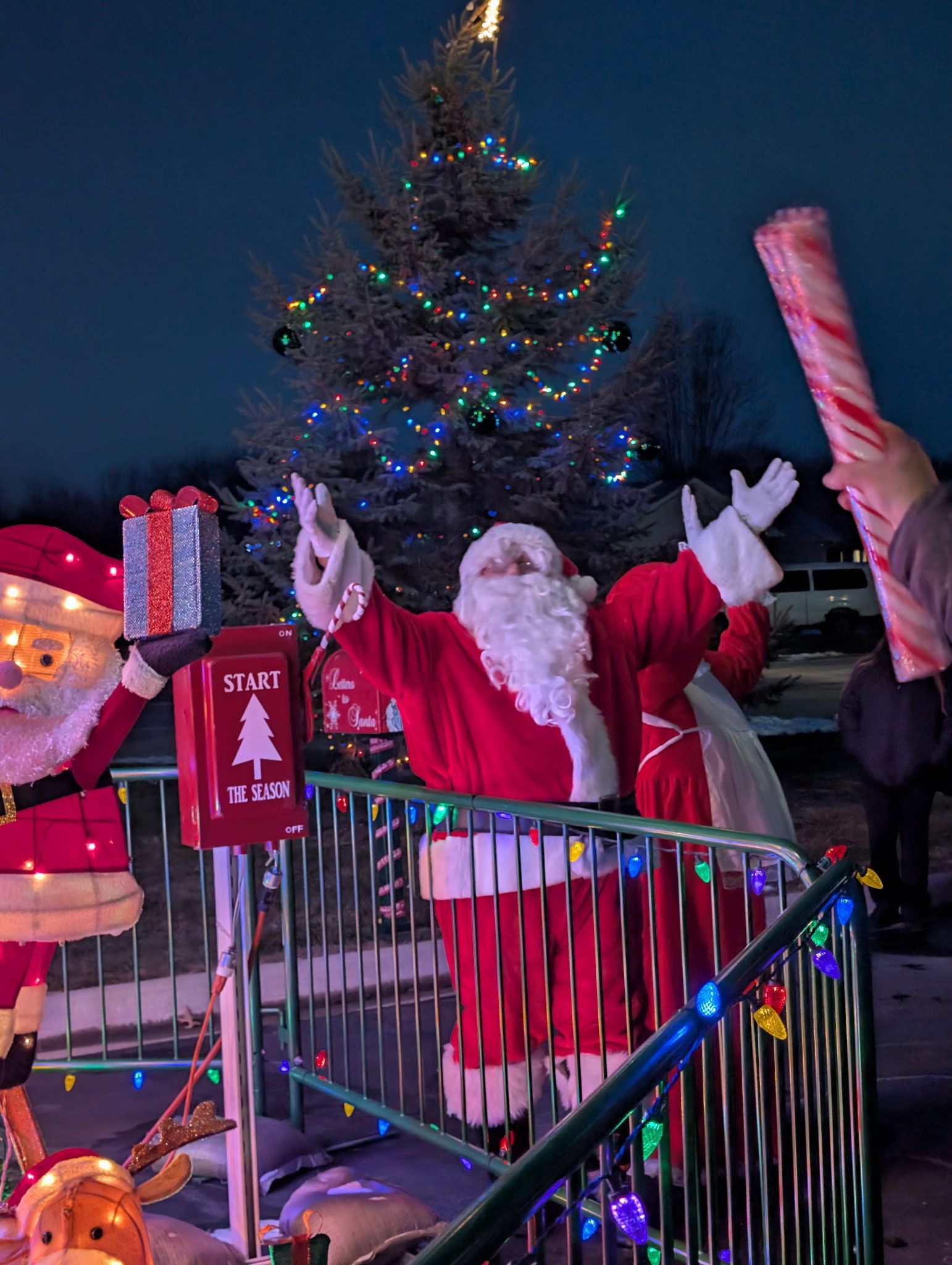 Santa waving at a festive holiday display with candy canes and colorful lights at night