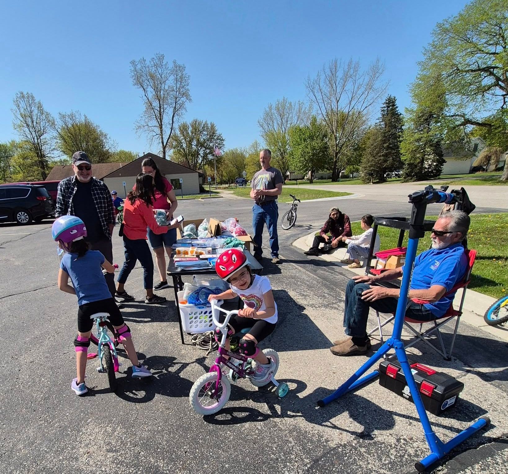 Family gathering outdoors with children on bikes, adults standing nearby, and a clear blue sky.