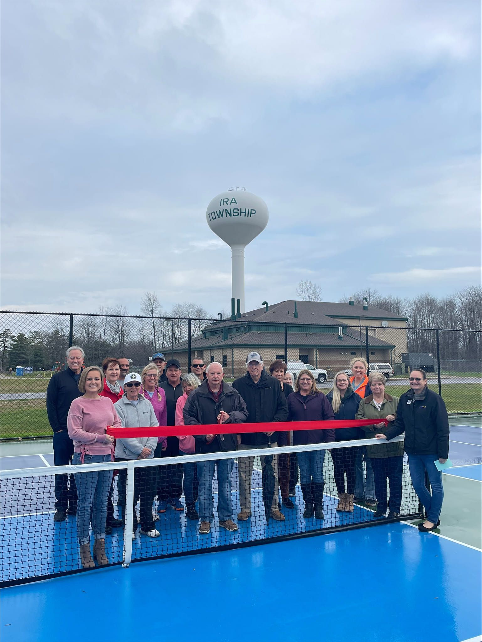 Group of people standing on a blue deck by a red ribbon, with a water tower in the background.