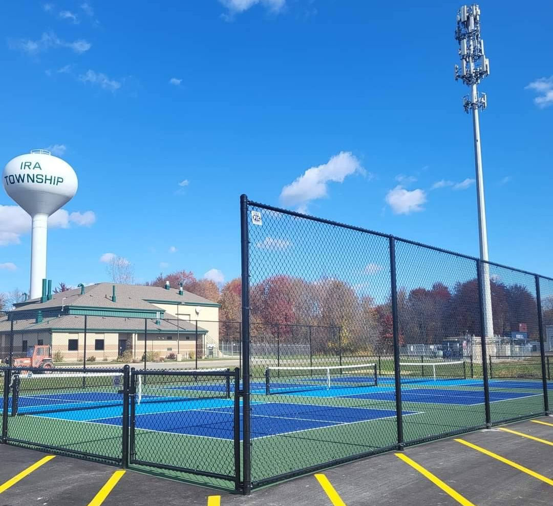 Outdoor tennis courts behind a black fence under a blue sky, with a water tower and light pole nearby.