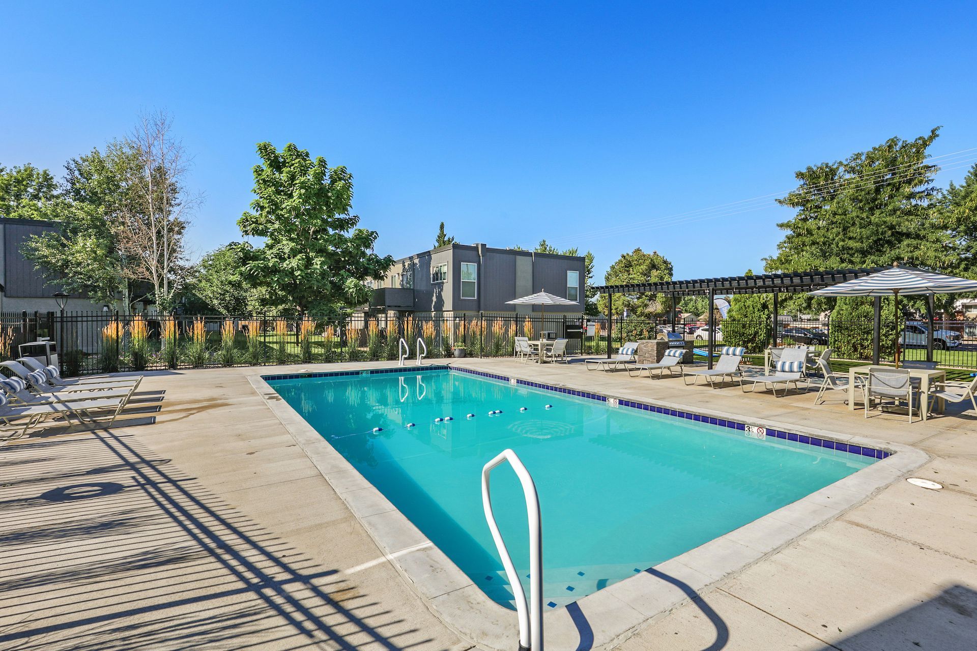 A rectangular swimming pool surrounded by a concrete deck, lounge chairs, and a pergola under a clear blue sky.