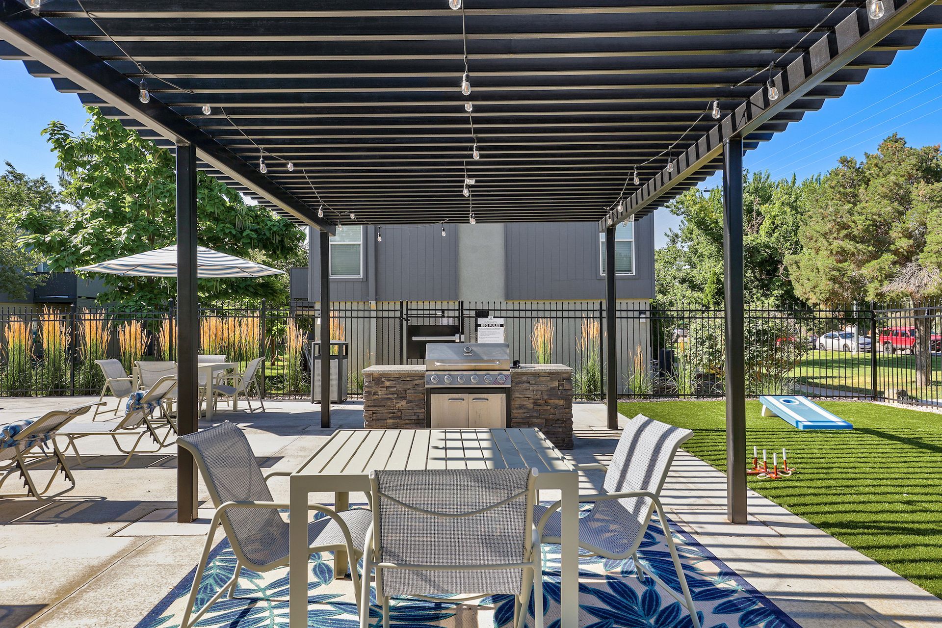 A covered patio dining area with chairs, a table, and a stone grill, set on a sunny day next to a grassy lawn.