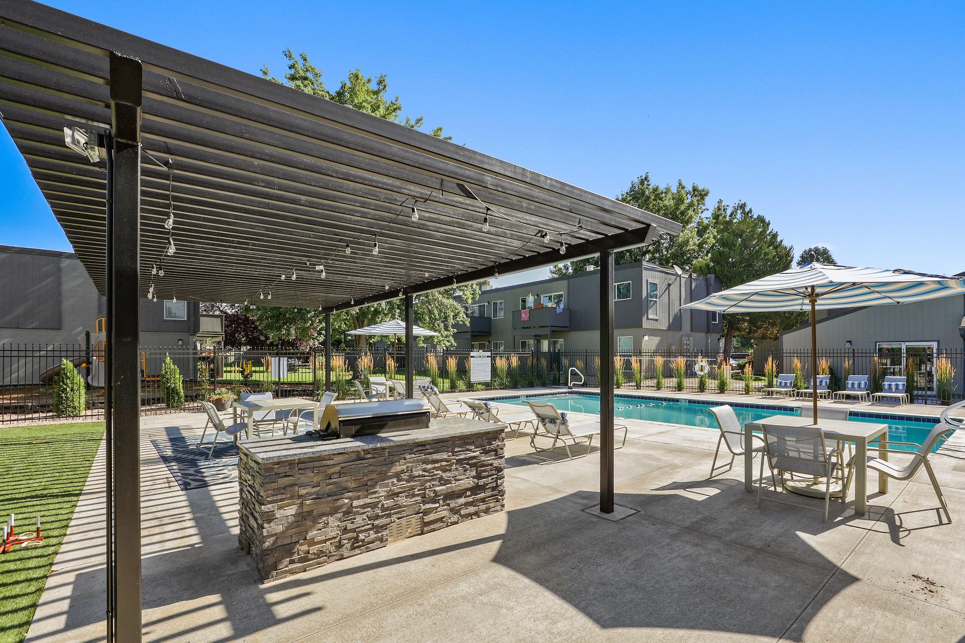 Outdoor swimming pool area with a stone grill island under a pergola, patio seating, and a large umbrella on a sunny day.