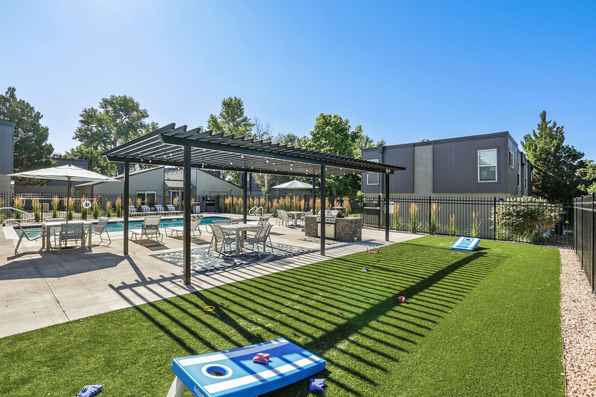 Outdoor apartment pool area with a pergola, seating, and a cornhole game on artificial grass under a clear blue sky.