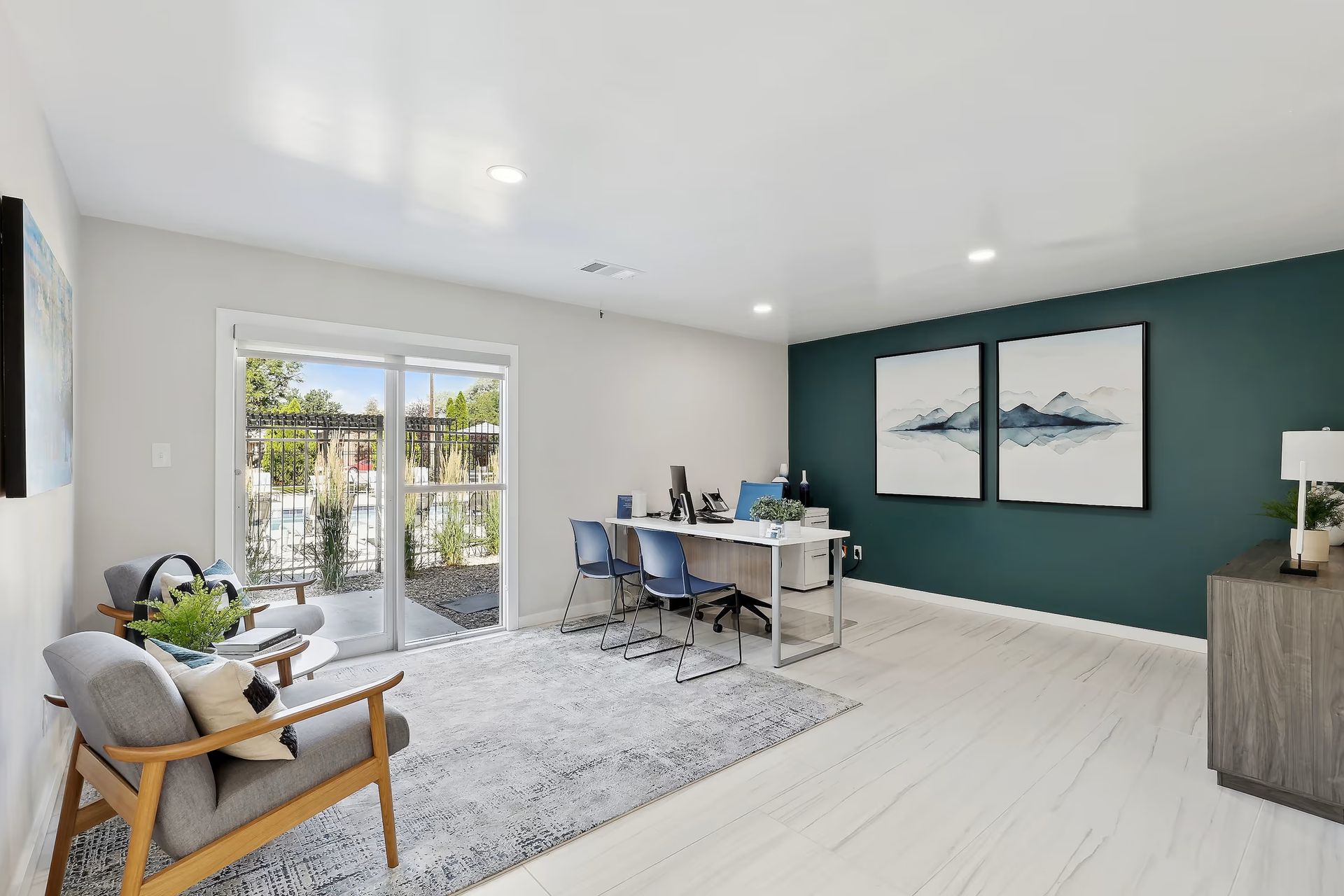 A bright home office with a desk, two blue chairs, a patterned rug, a grey armchair, and wall art against a teal wall.