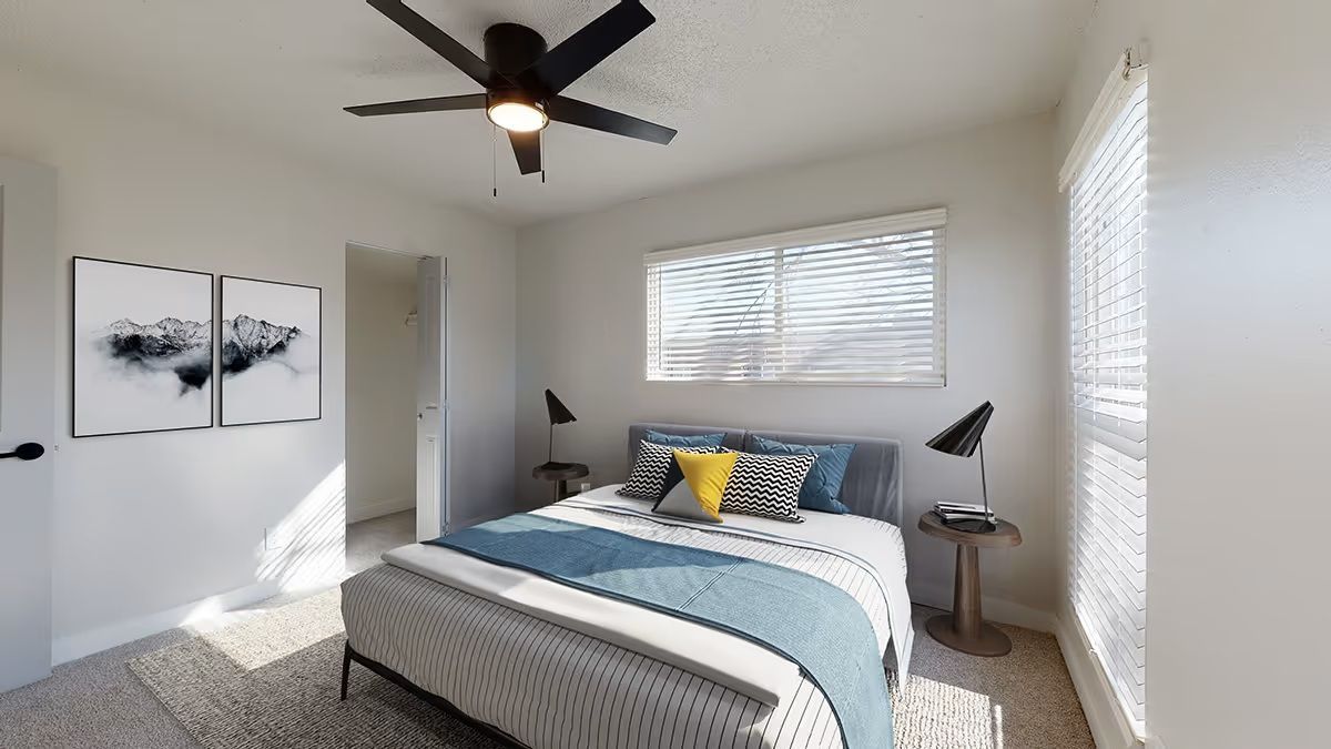 A modern bedroom featuring a bed with blue accents, two small side tables with lamps, a ceiling fan, and abstract art.