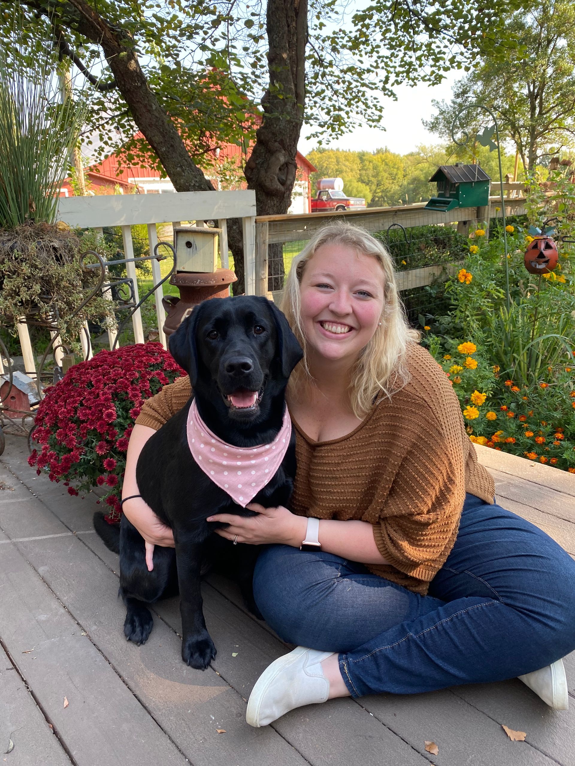 Woman sits cross-legged on a deck, smiling with a black dog wearing a pink bandana. Fall foliage in the background.