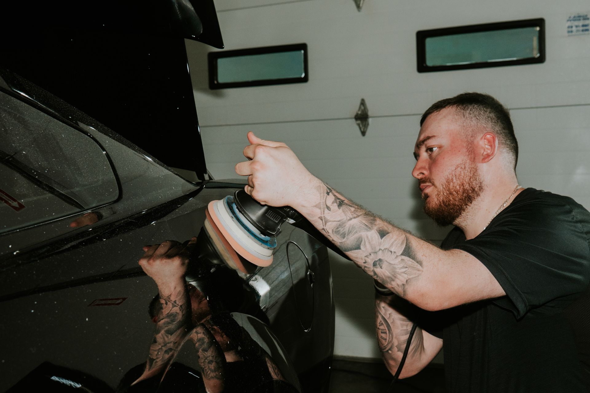 Man polishing a black car with a buffer in a garage, wearing a black shirt and tattoos visible