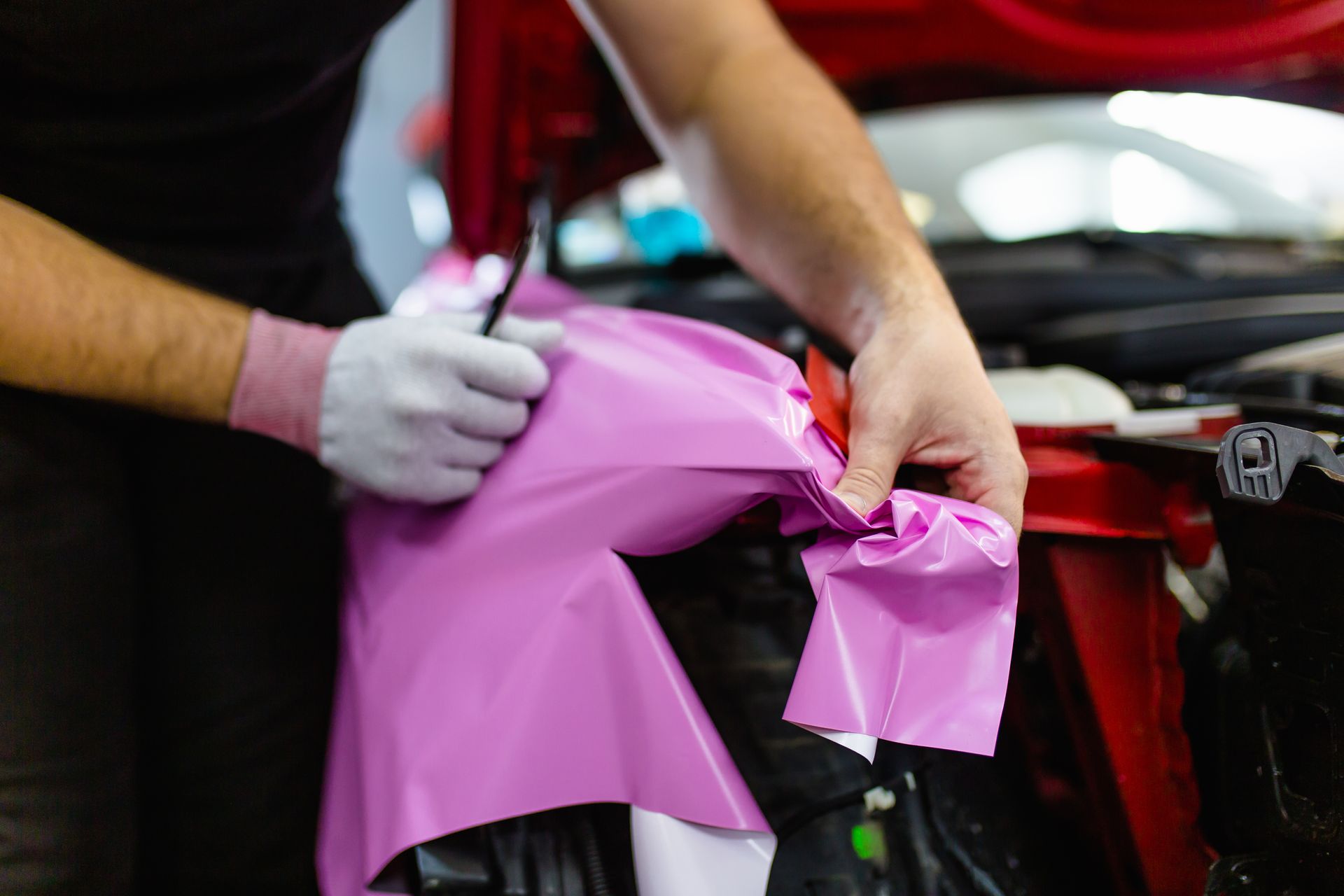 Hands applying pink vinyl wrap to a car panel in a workshop
