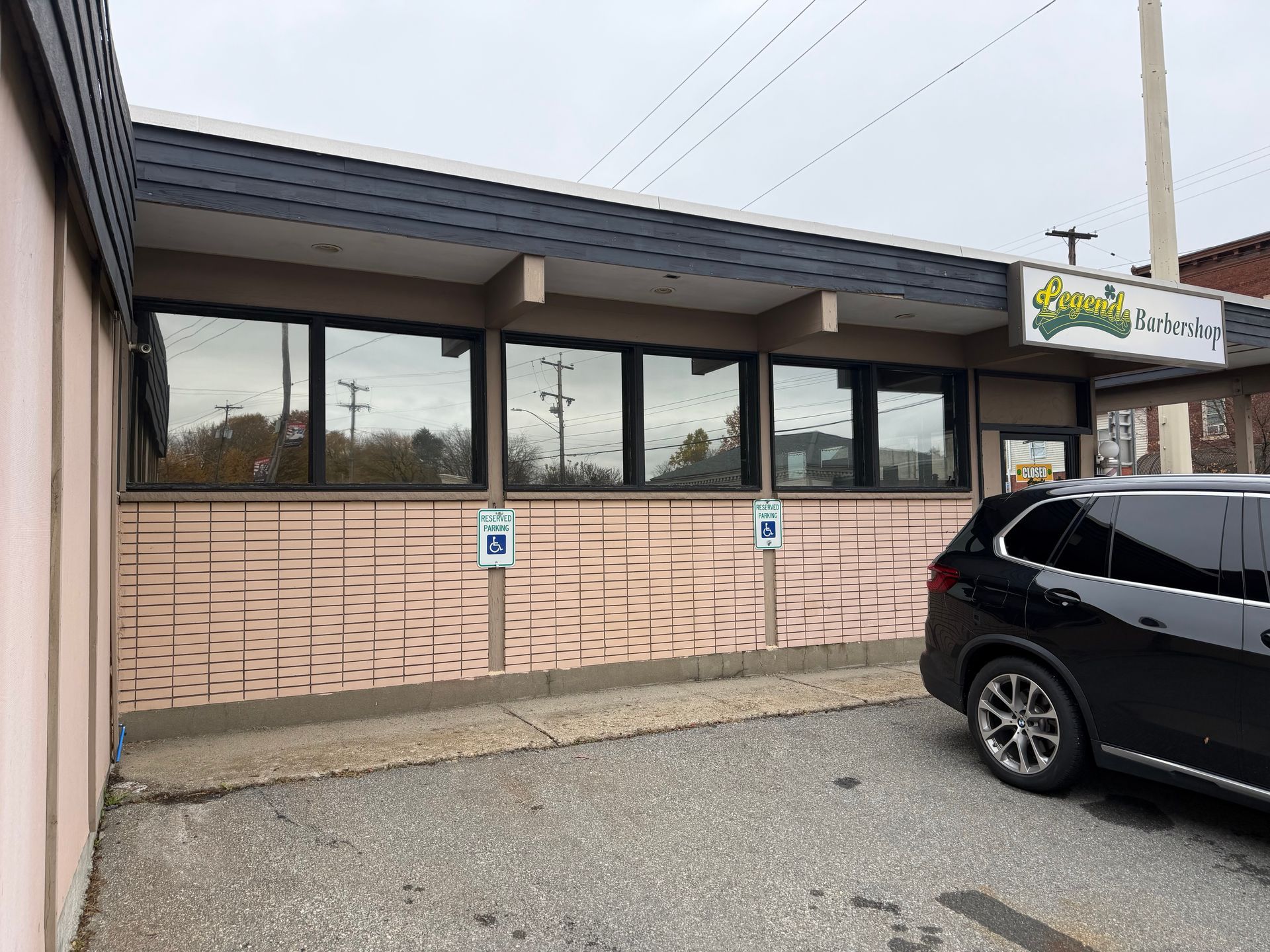 Strip mall storefront with dark windows, tan brick facade, and a parked black SUV in front