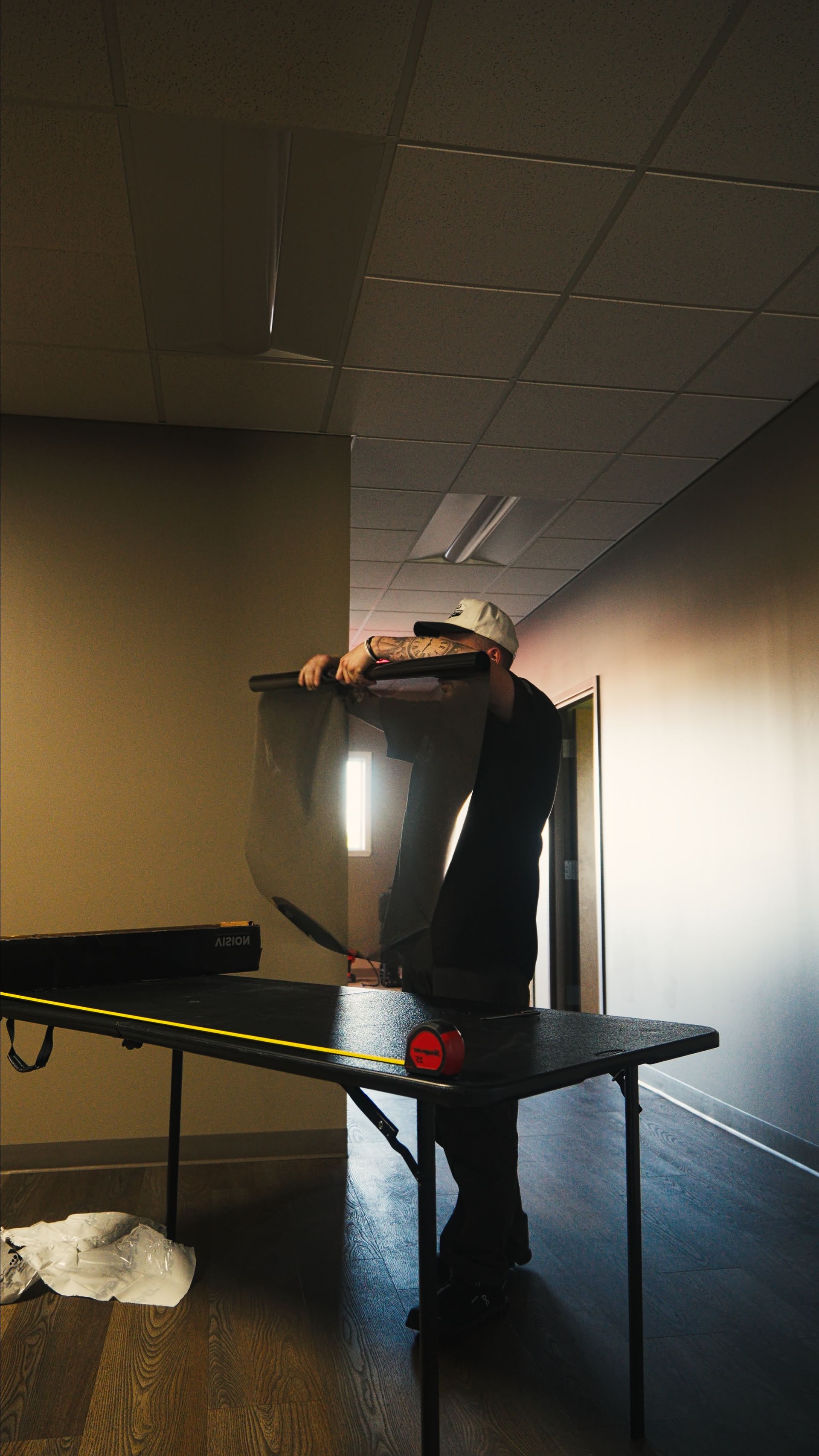Person standing on a table in a dim hallway, reaching up near the ceiling with a bright light behind them.