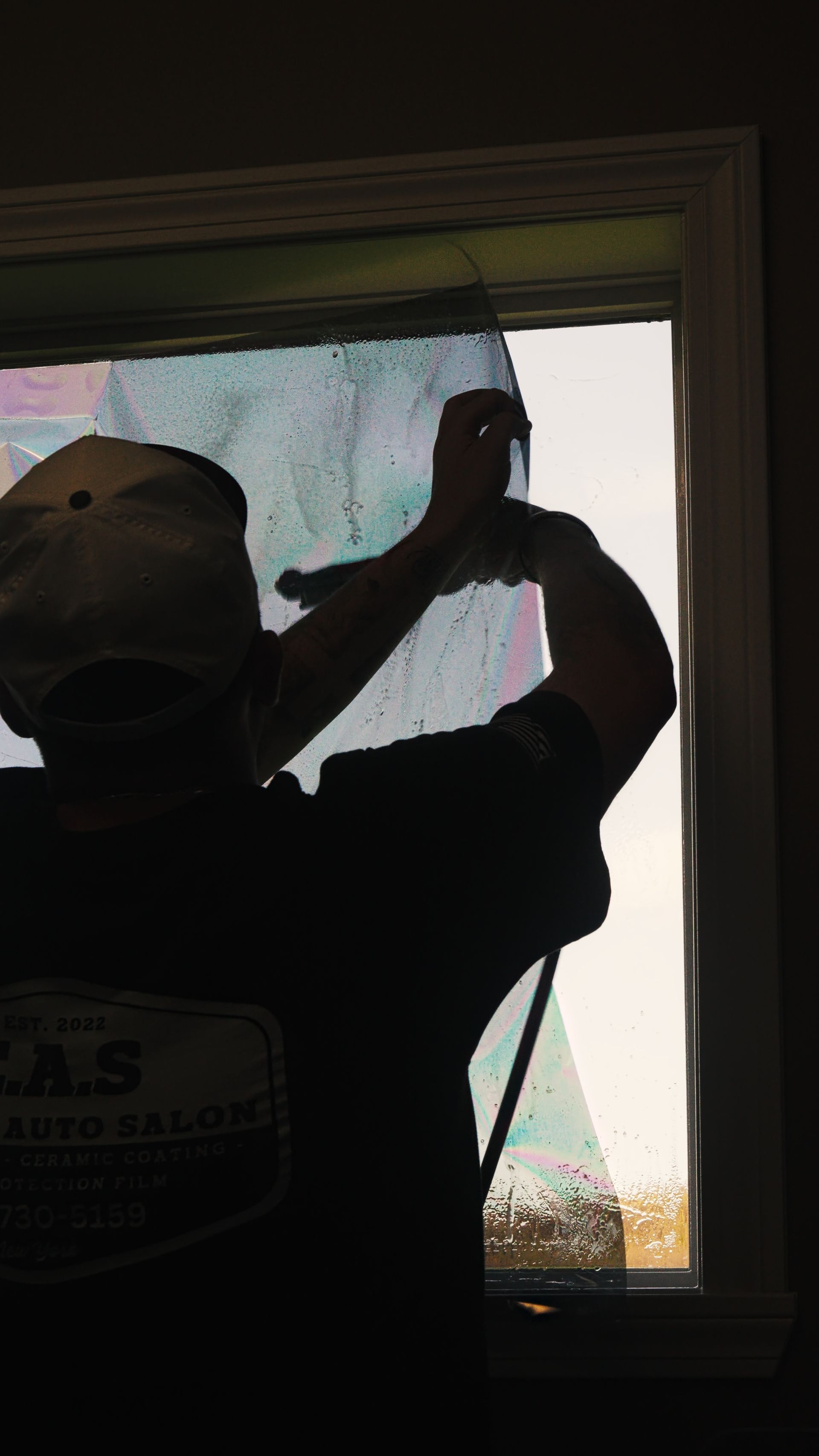 Person silhouetted at a window, lifting a tool to scrape or paint the glass frame.
