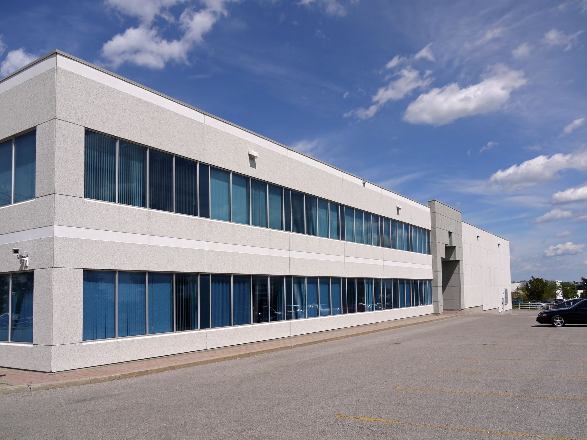 Modern white office building with blue-tinted windows and a large empty parking lot under a partly cloudy sky