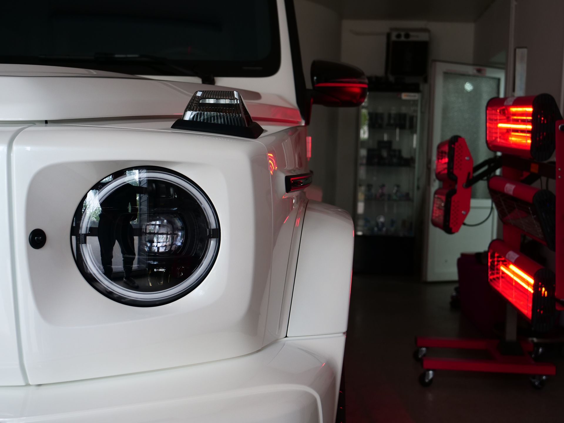 White SUV rear with round tail light, next to a red-lit vehicle in a garage or showroom