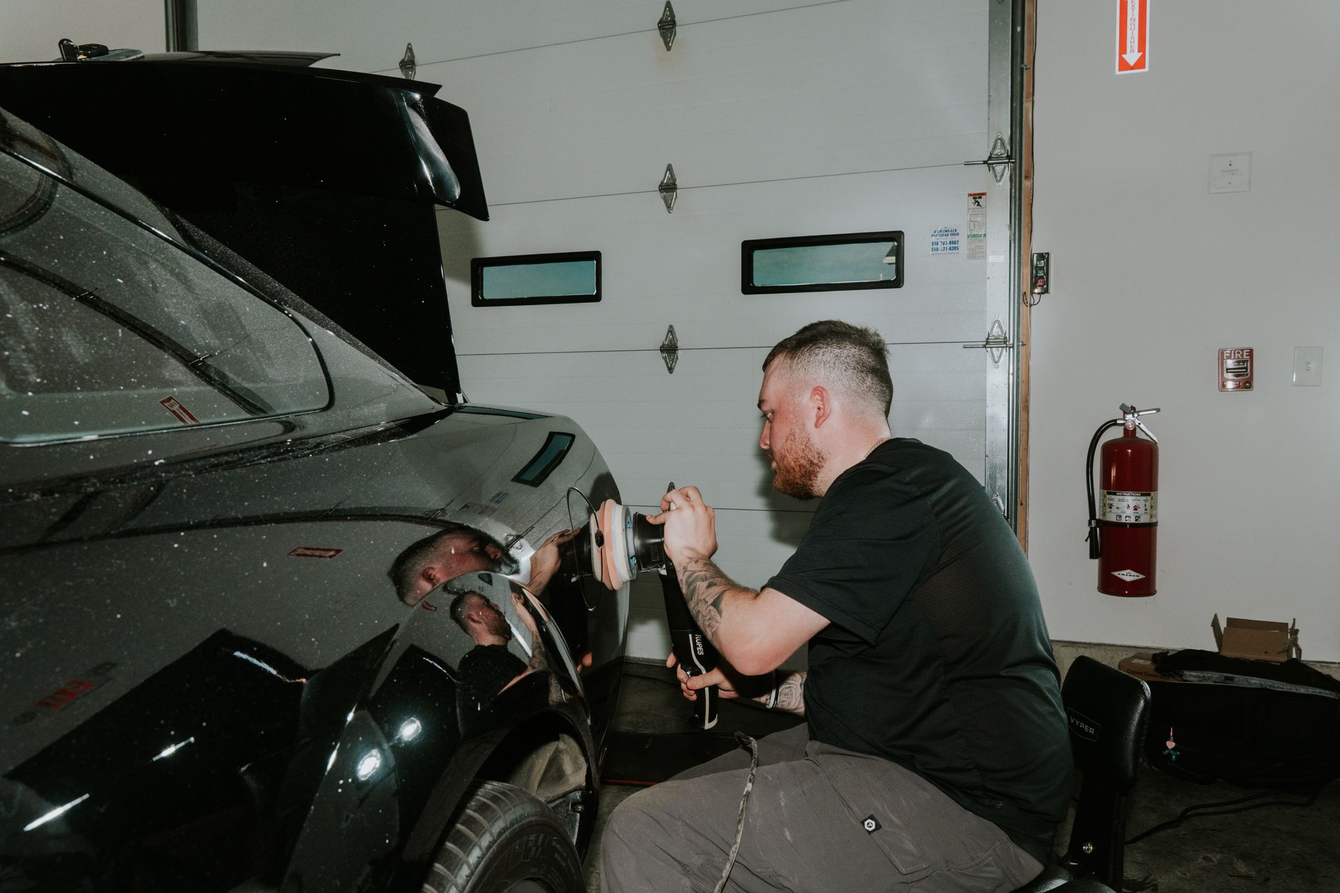 Mechanic polishing a black car in a garage beside a white door and fire extinguisher